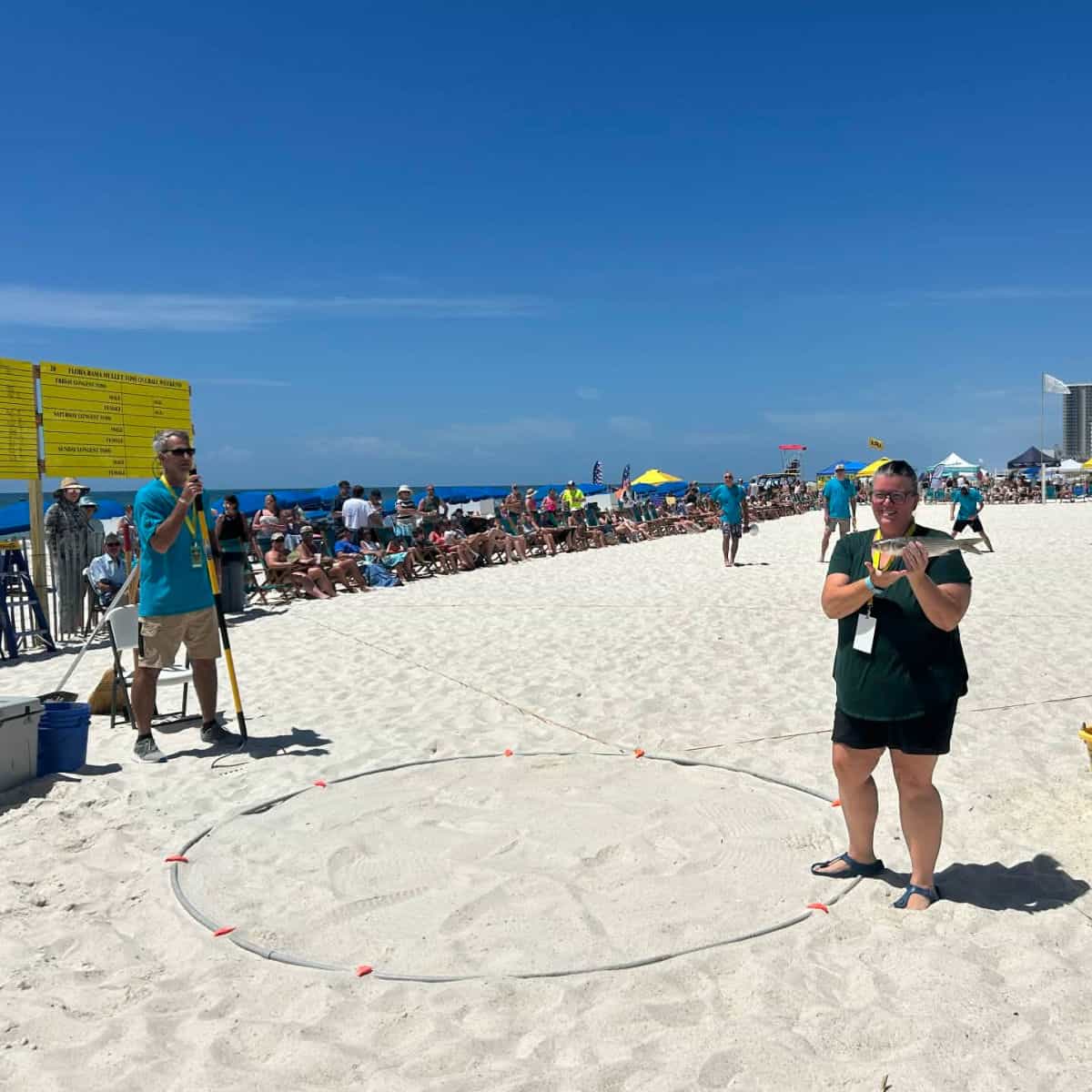 Tammilee holding a Mullet fish, standing in a circle in the sand, people lining the side of the beach for Mullet Toss at the Flora Bama