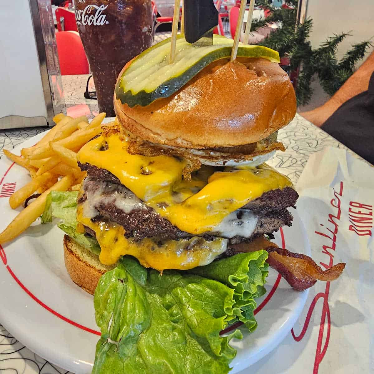 Jukebox Burger at Sunliner with three burger patties, melted cheese, pickle on top of the bun, fries on a white plate with red circle next to a paper with Sunliner Diner printed on it and a glass of Coke