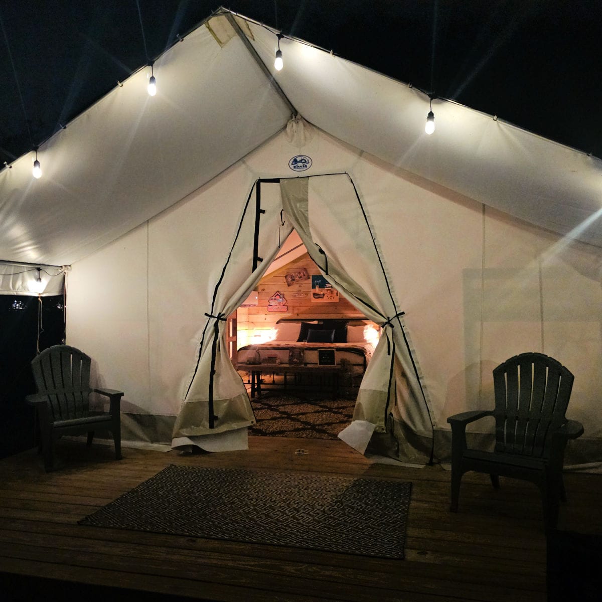 timberline glamping tent in Orange Beach at night with the lights on inside the tent and a bed seen inside the tent, two Adirondack chairs on the front of the tent.