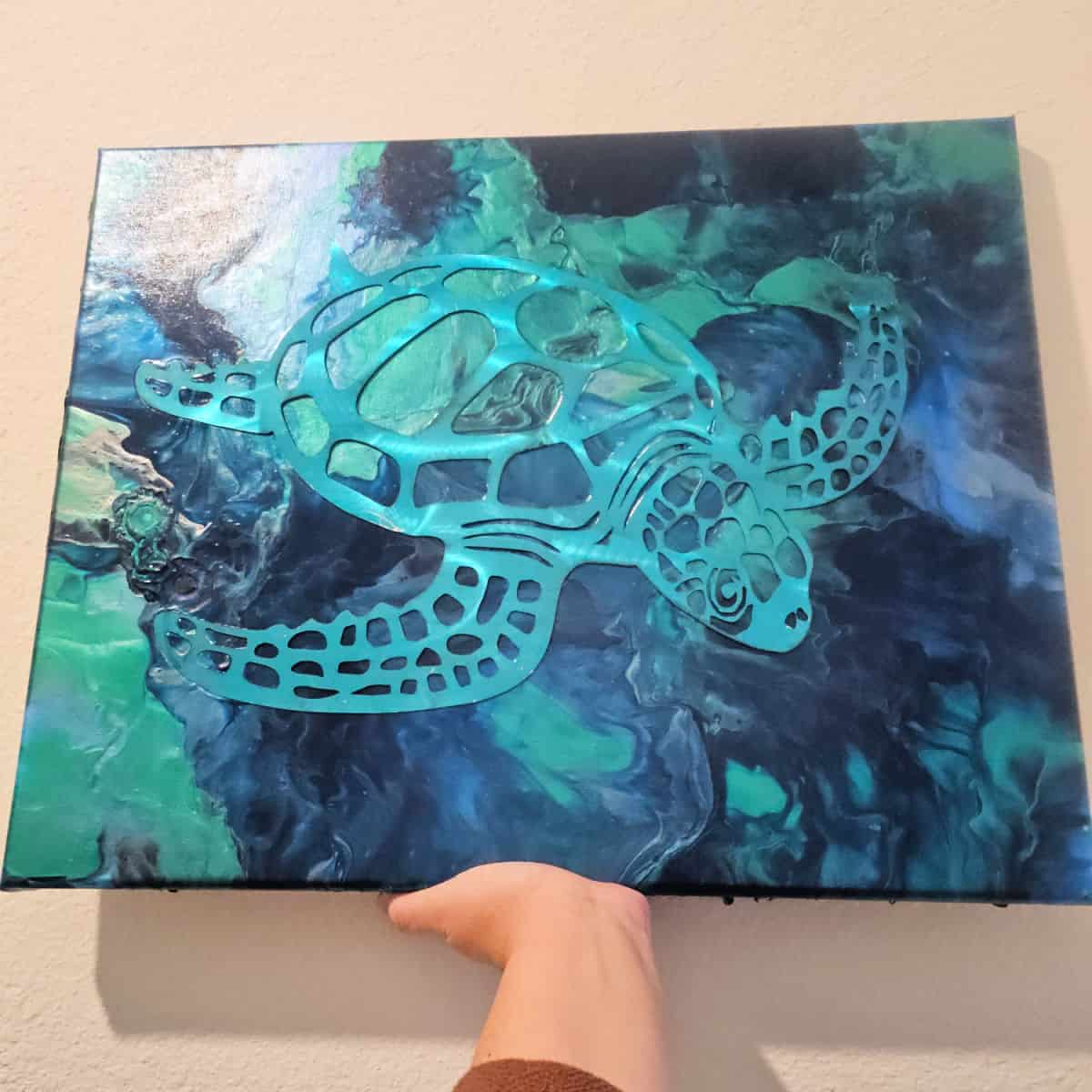 hand holding a fun turtle painting with blue and green backgrounds and a metal turtle on it against the wall