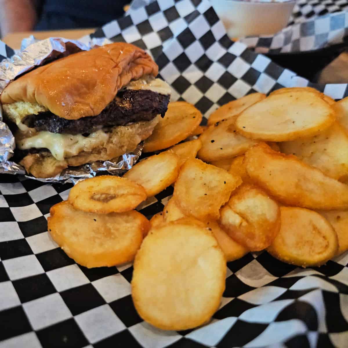 burger wrapped in foil sitting on a checkered paper lined basket with potato medallions