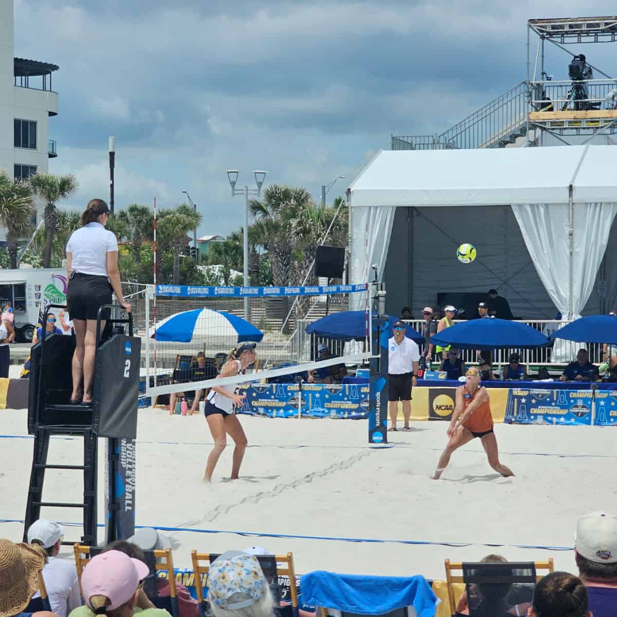 NCAA beach volleyball match wtih two players seen on the sand and a ref on a tall ladder, cameras in the background above a tent