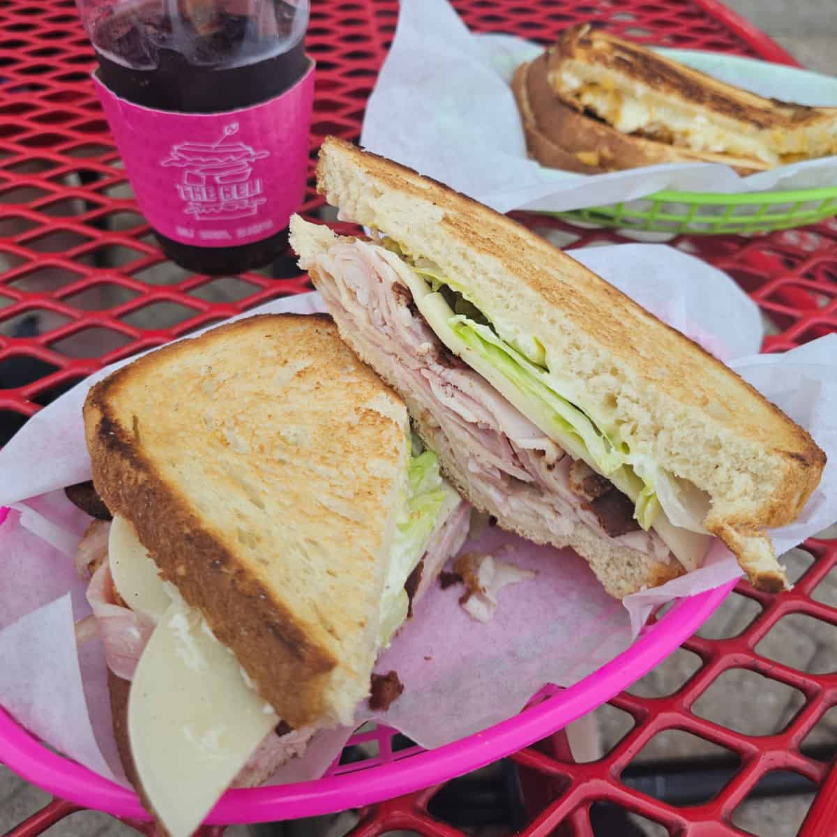 sandwich with meat, cheese, and lettuce in a paper lined basket next to a beli deli drink and a green basket with a sandwich