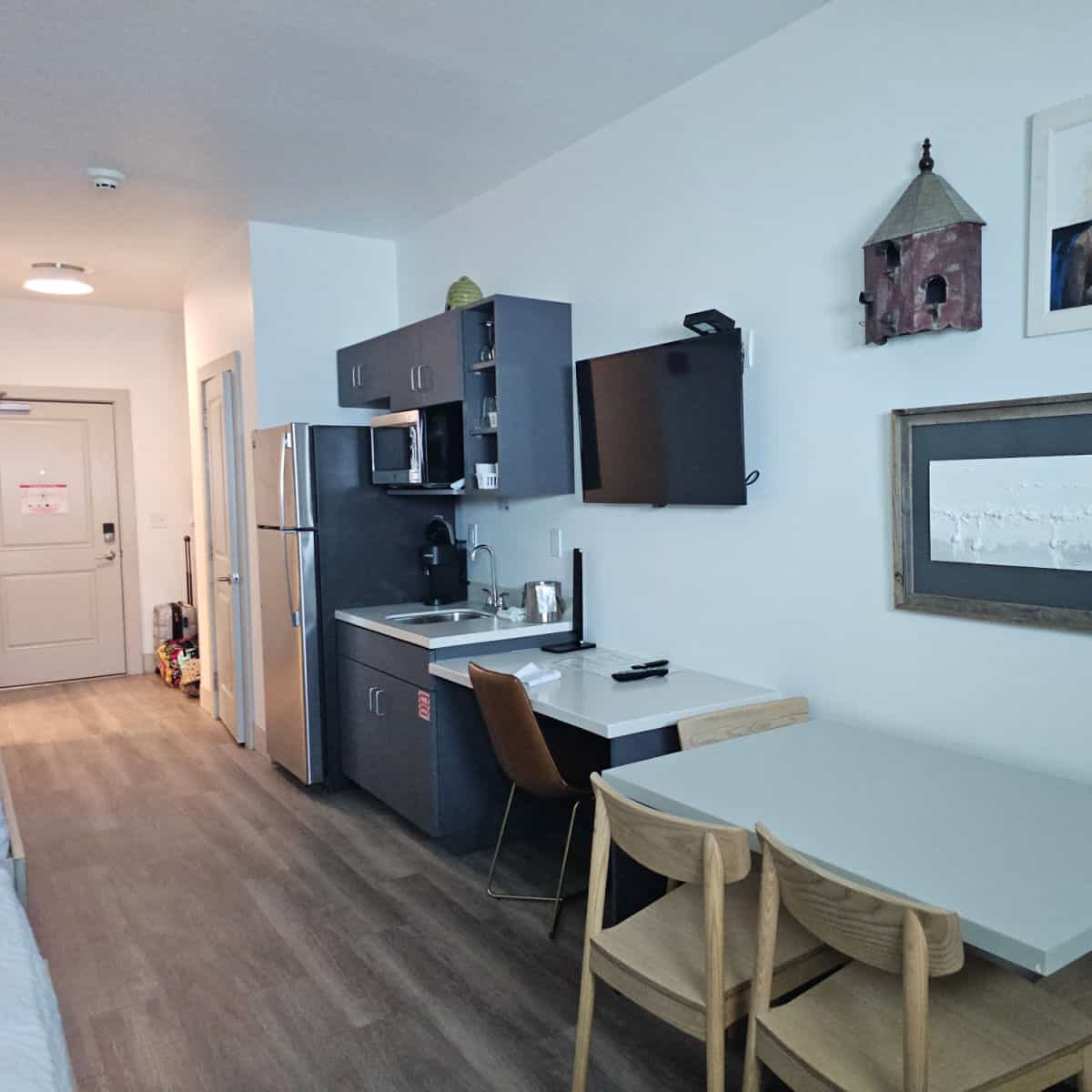 hotel room kitchenette area with full size fridge, sink and counter, microwave next to cabinets, flatscreen tv, bird house, and art on the wall above a table and chairs
