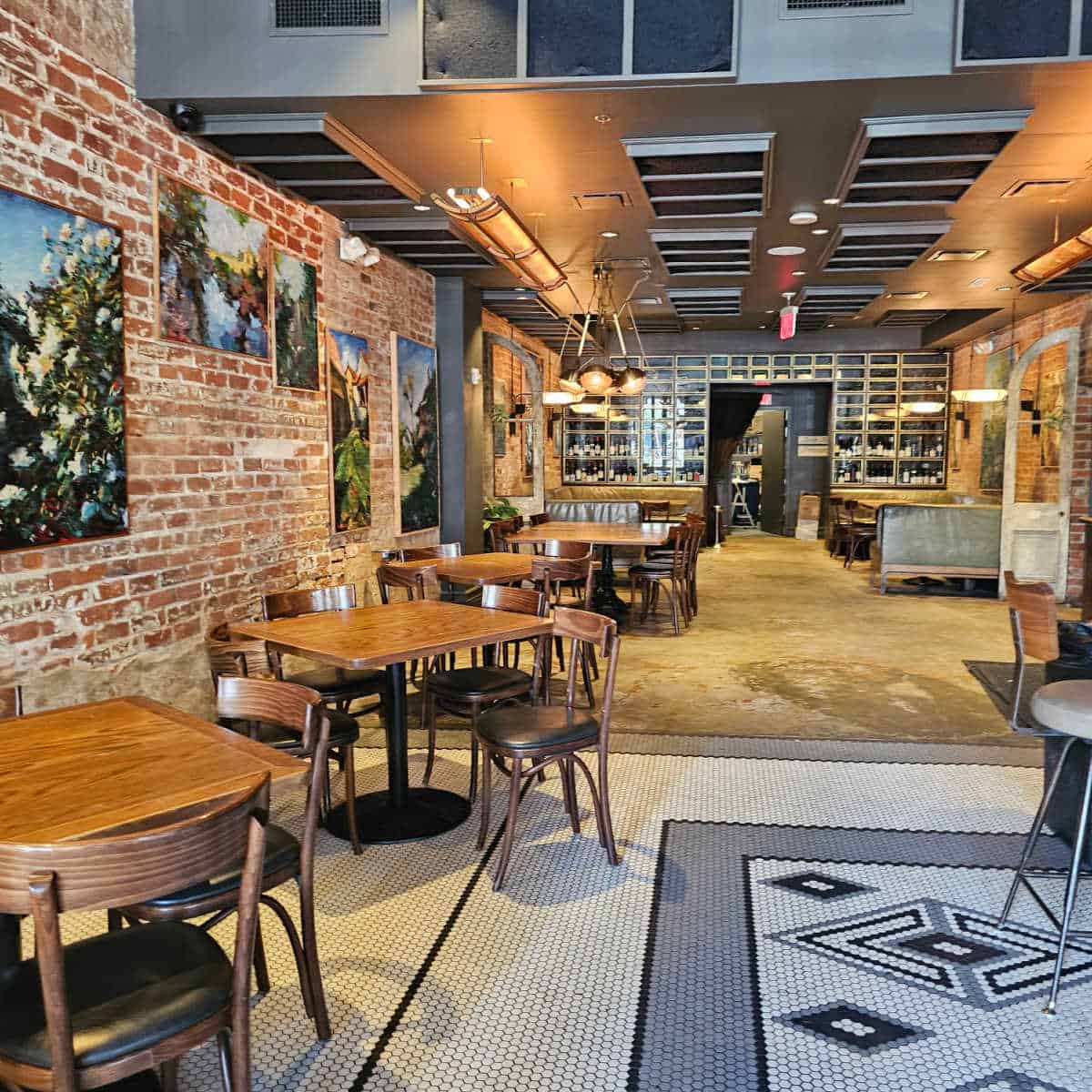 Compare Lapin dining area with tiled floor, wood tables with chairs, brick wall, and a wall of wine in the background
