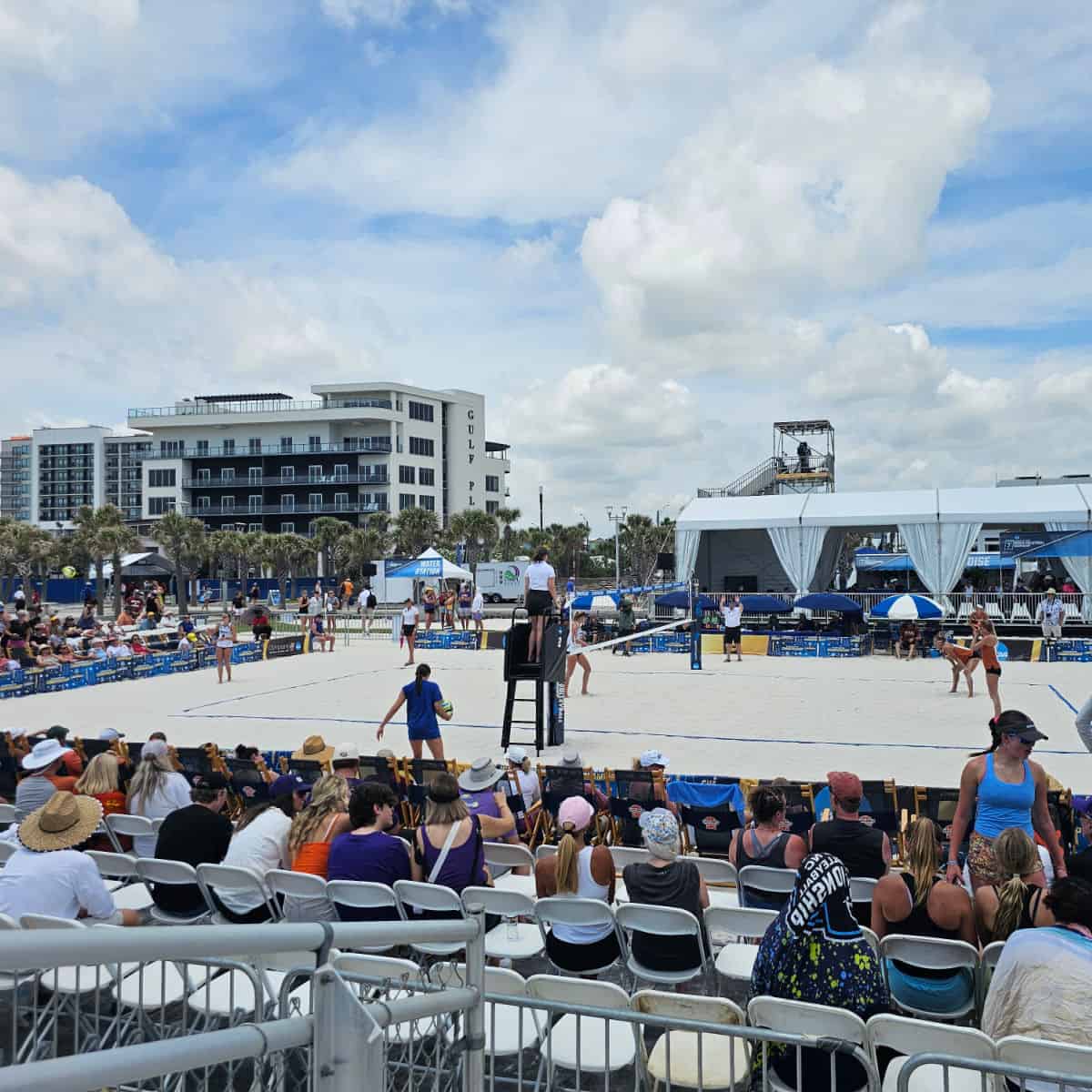Looking over chairs to a beach volleyball match, white tent in the background with camera stand above the tent