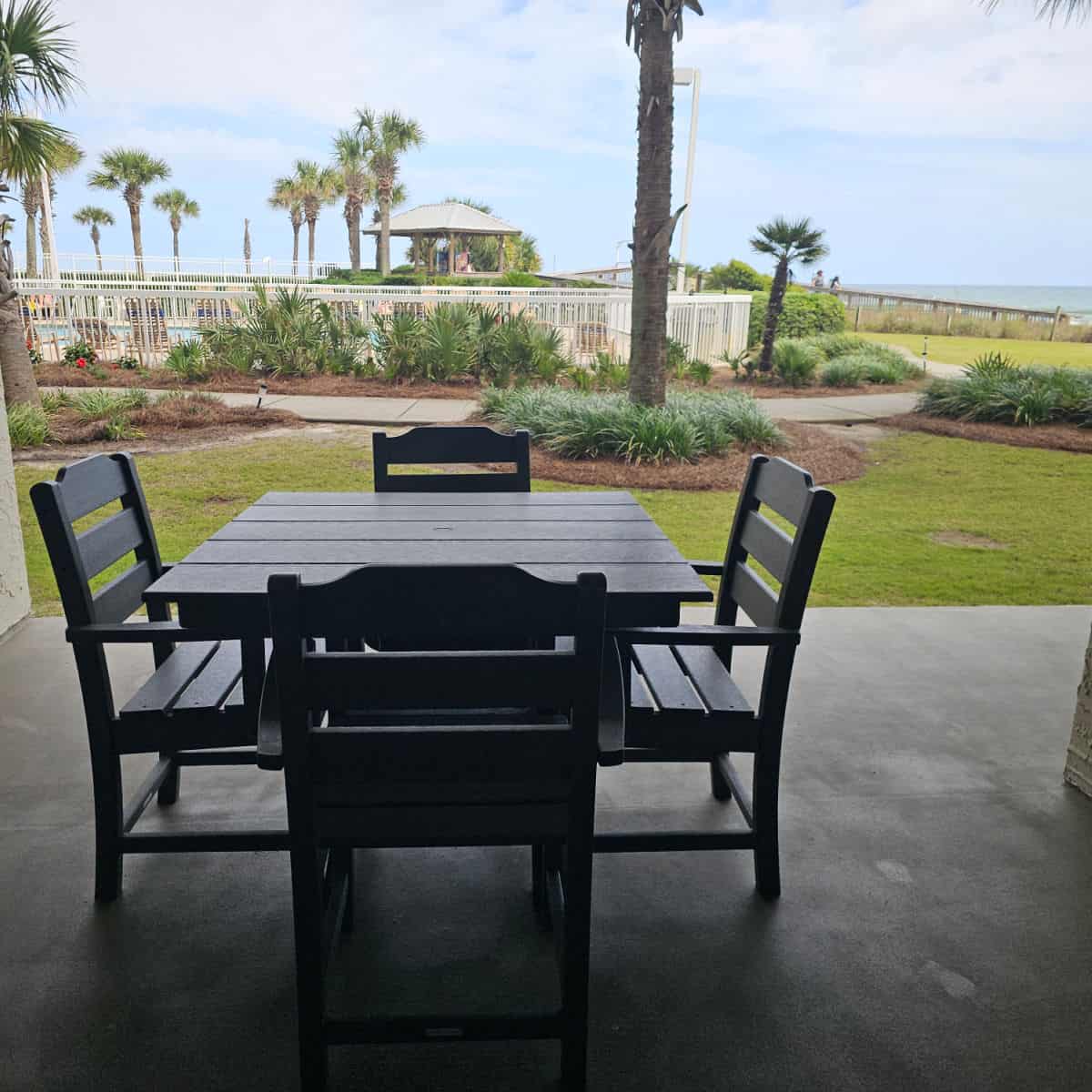 Table and chairs on a paved area, palm trees in the background with fence around a pool