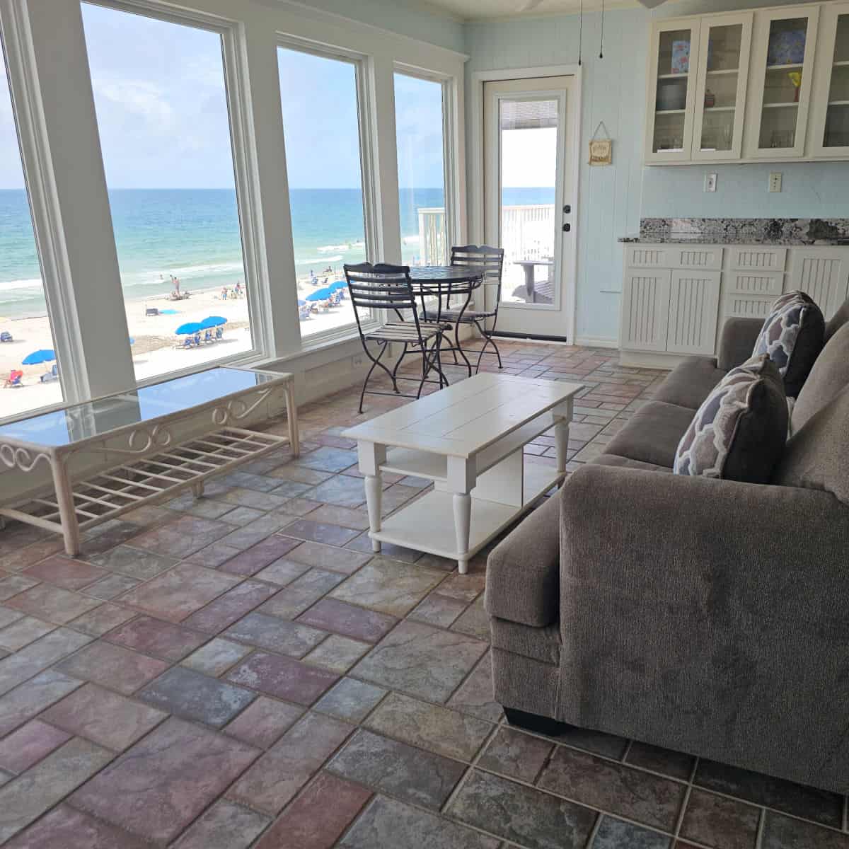 all weather room with windows looking out to the beach, grey couch with pillows and a white table.