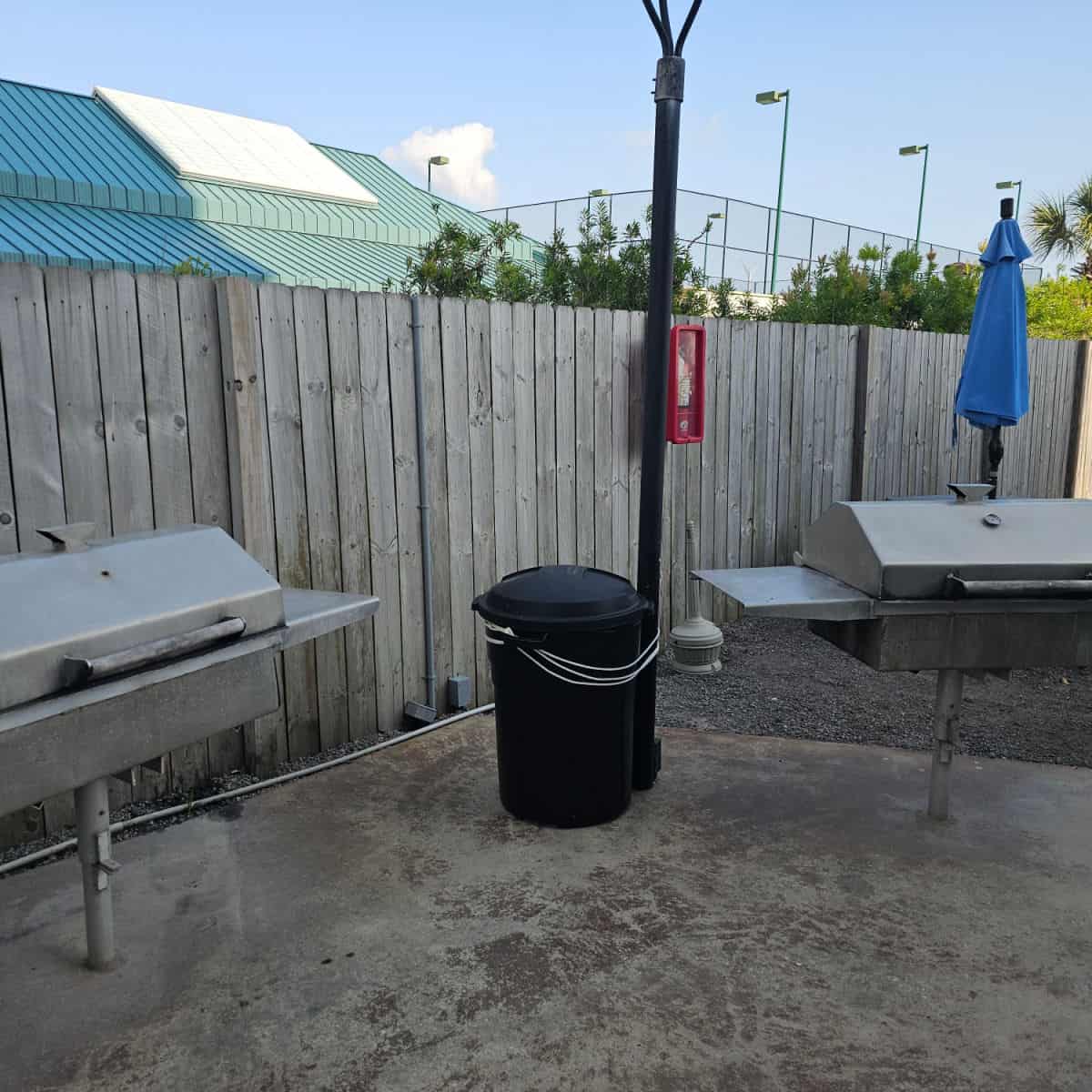 two barbecues with a garbage can in between them, next to a fence
