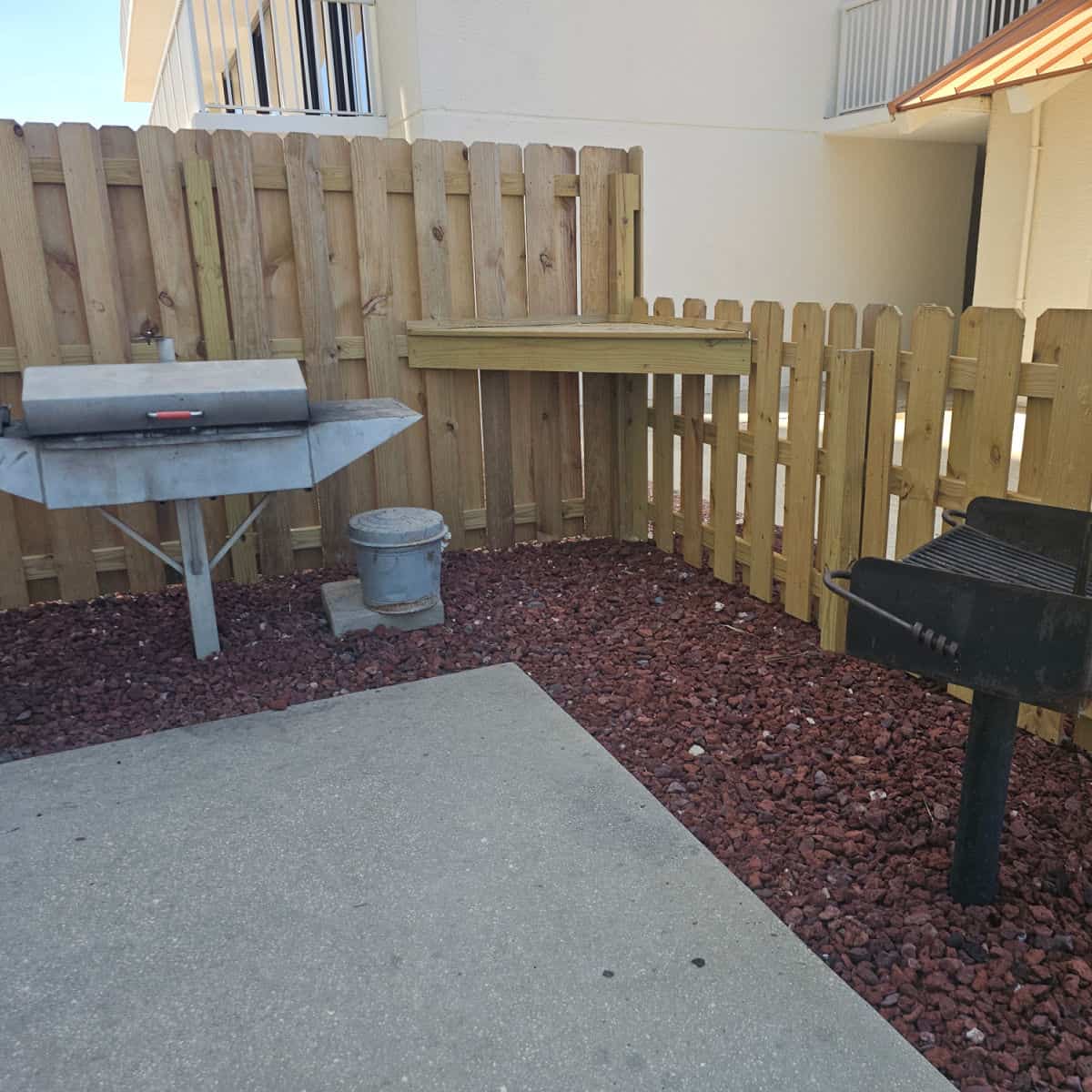 barbecue are with two different barbecues and a metal garbage can, fence along the back
