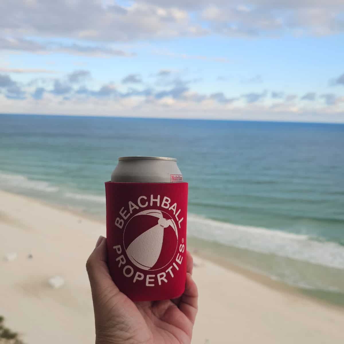 hand holding a can in a beachball properties coozie with the gulf and beach in the background
