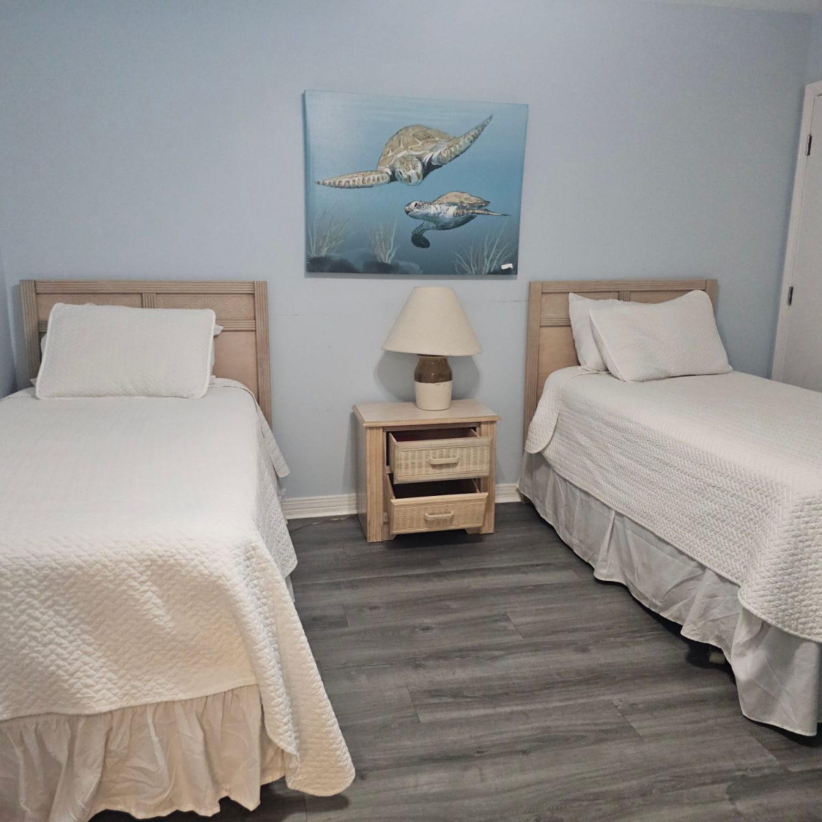Two twin beds with white linens, table and lamp in the middle of the beds, turtle art on the wall