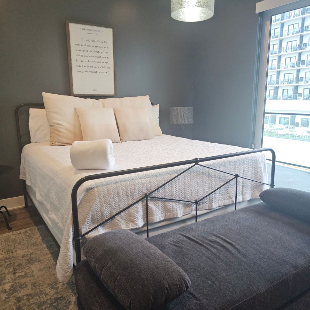 bedroom with large bed, white linens, grey couch at the end of the bed, print on the wall, looking out the window to a hotel across the street