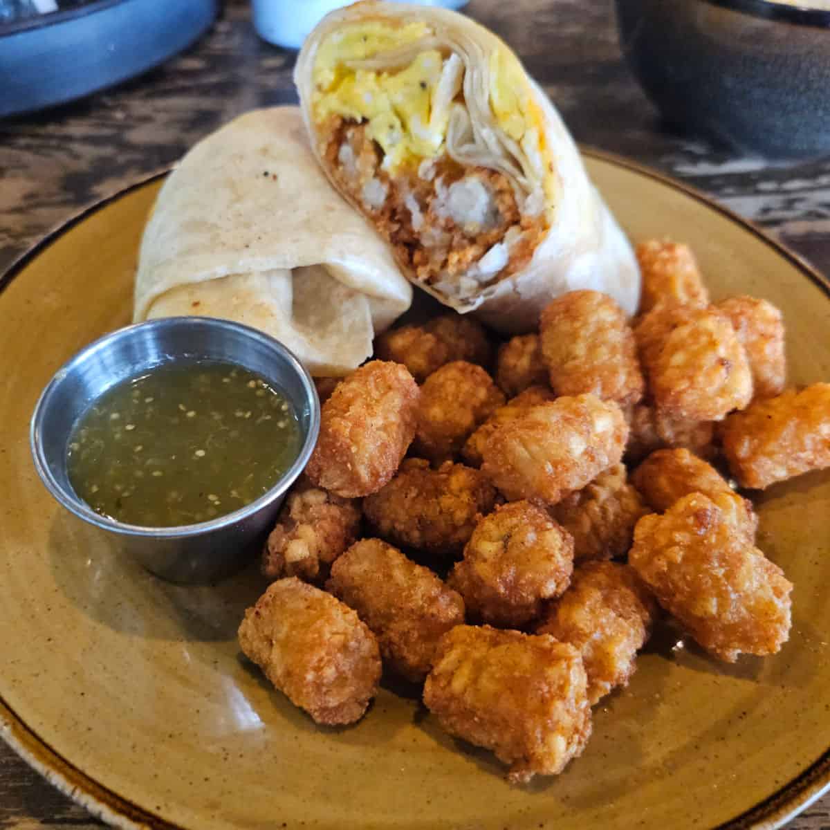 beakfast burrito on a yellow plate with tater tots and green sauce in a container
