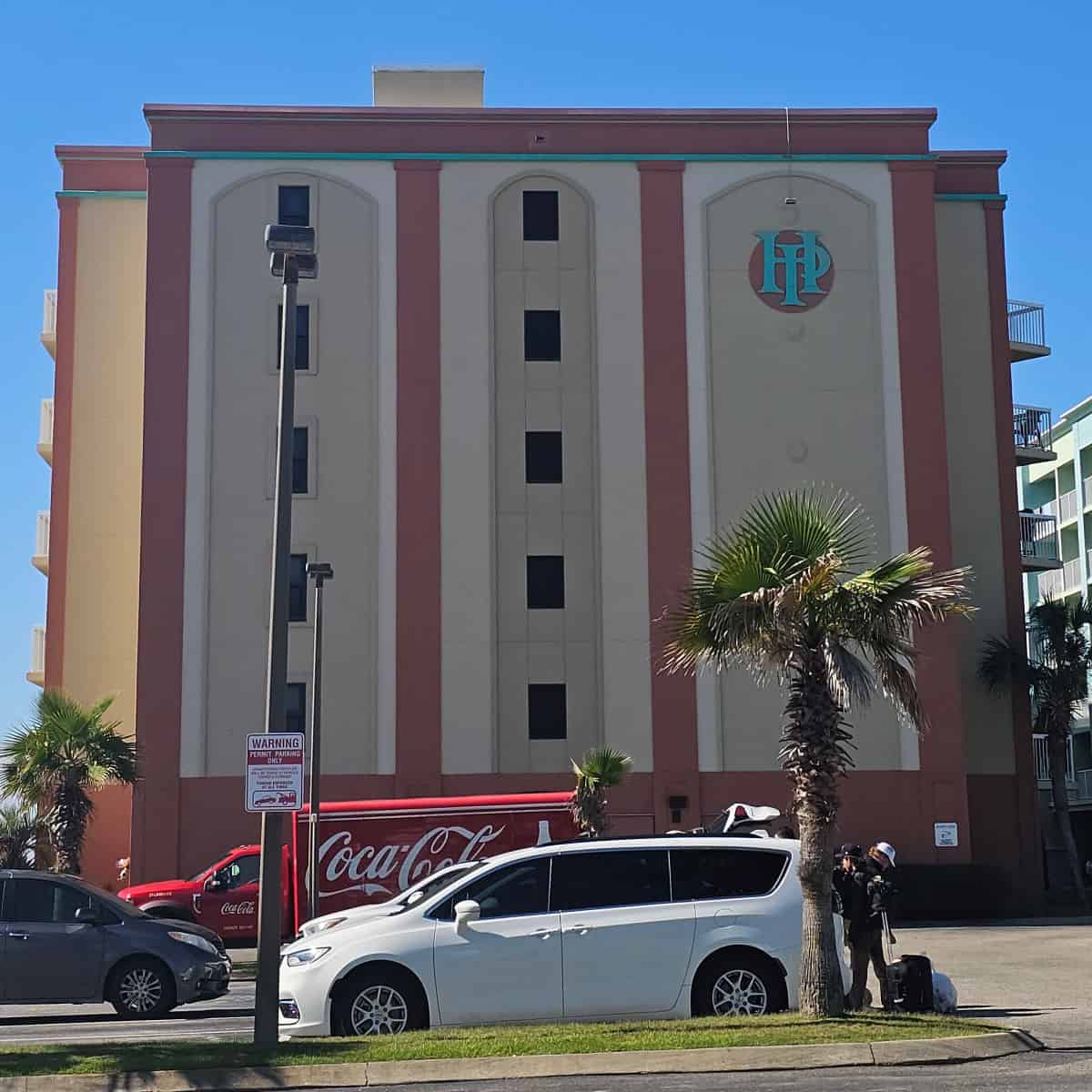 HP in turquoise on the side of a pink and cream colored building, cars and a COKE truck in the parking lot near palm trees