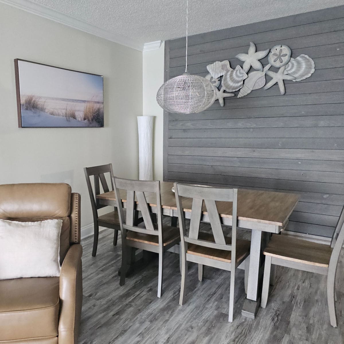 dining room area with grey shiplap wall, shells on the wall, dining room table and chairs next to a brown leather chair