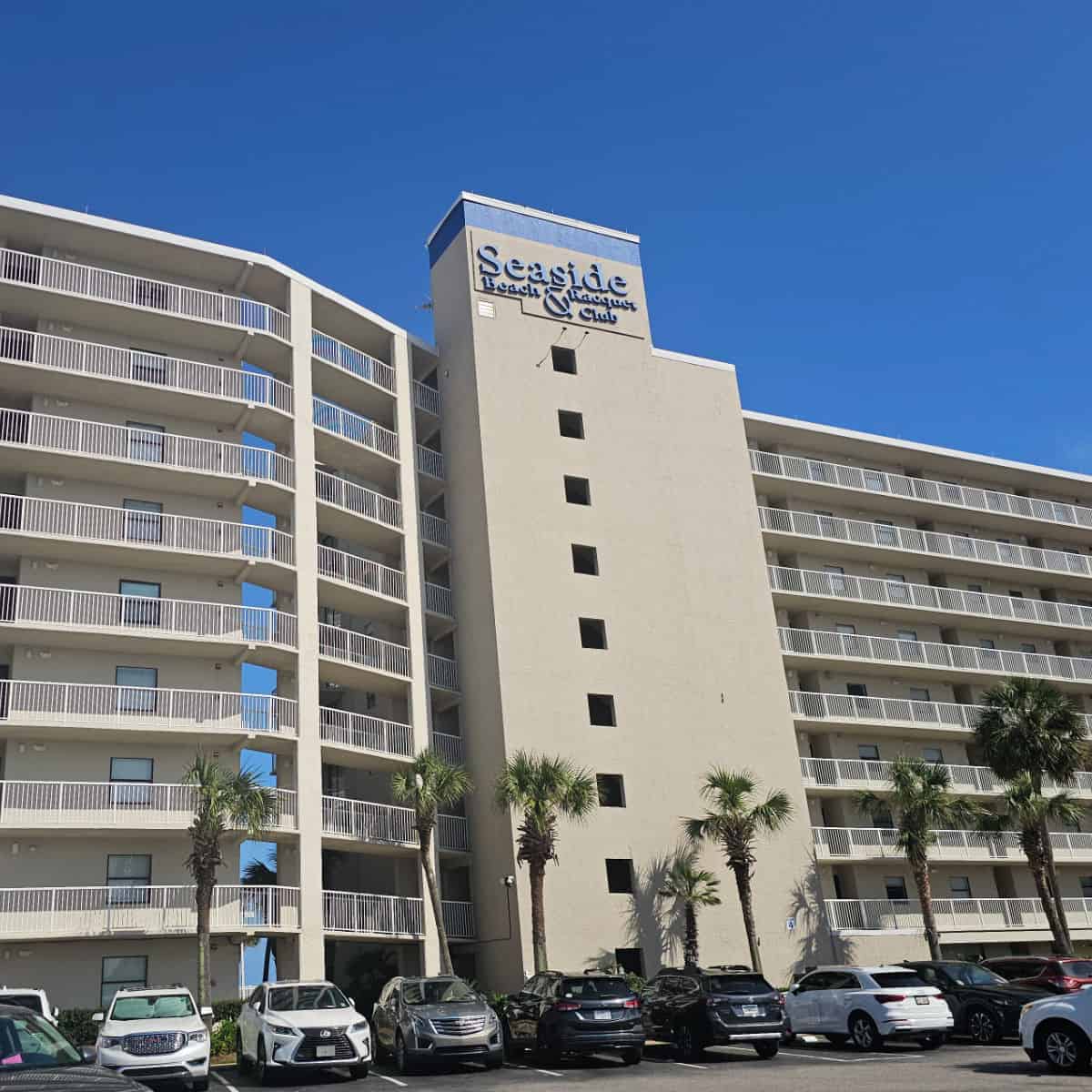Seaside Beach & Racquet Club sign on the side of a large condo building, cars in the park area