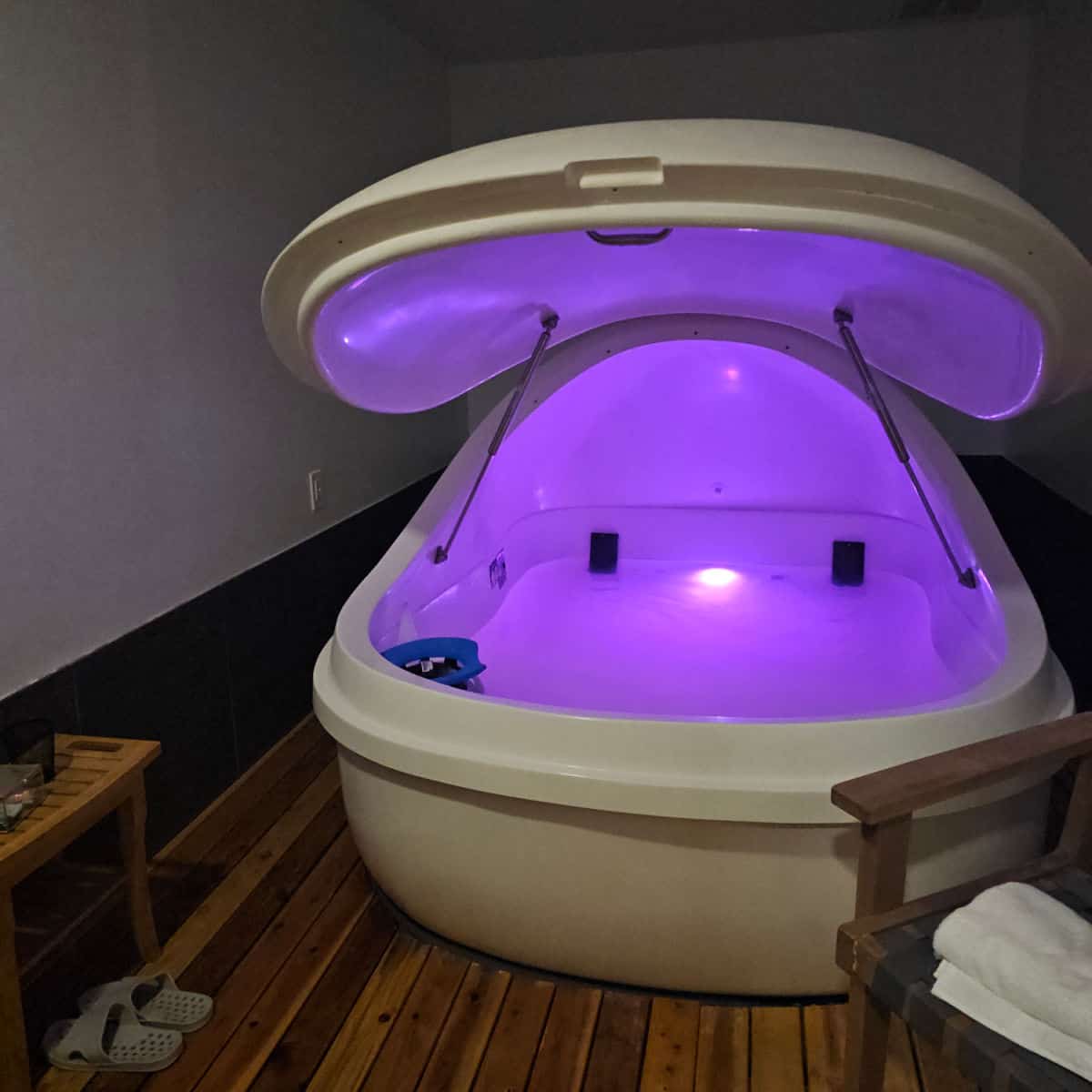 Float therapy pod with purple lights, a chair next to the pod, and table with plastic slippers on the floor