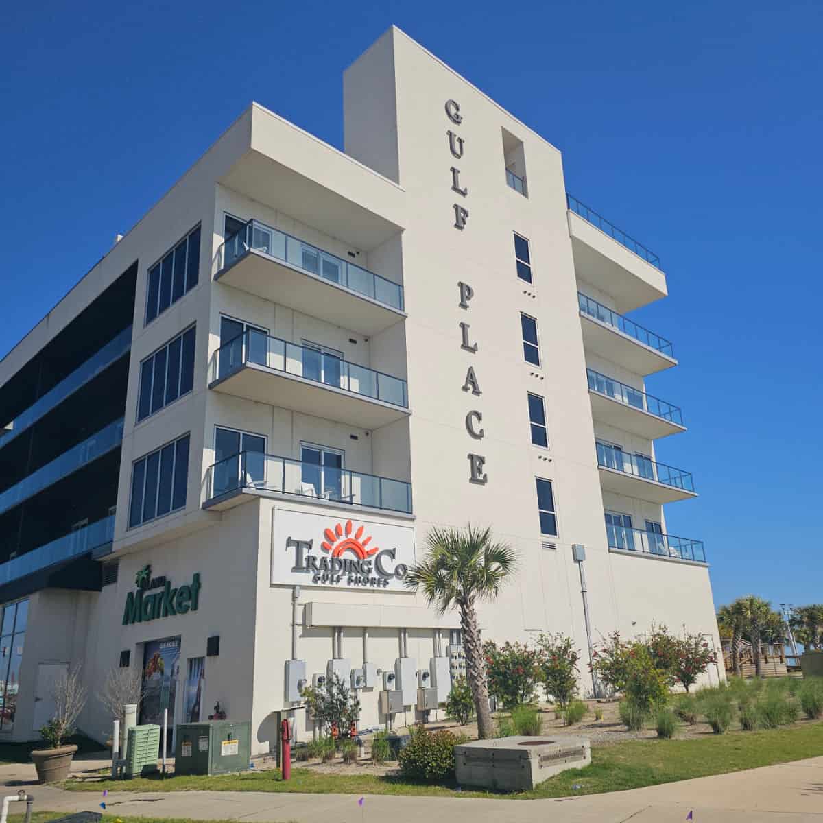 Gulf Place sign along the side of a multi-story building with Trading Co Gulf Shores on the base floor, palm trees and brush around the base