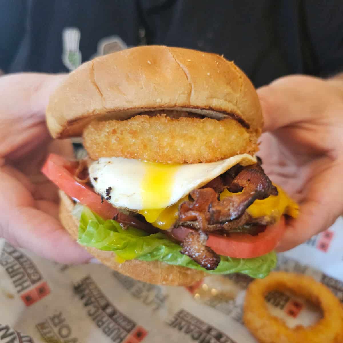 hands holding a burger with an onion ring and egg dripping down the side