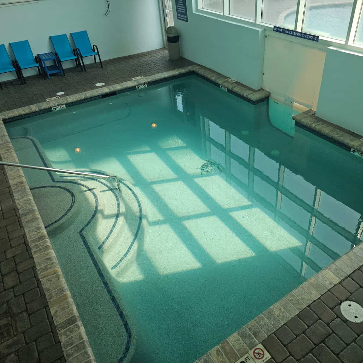 Indoor pool with steps leading down into the pool, blue deck chairs on the far wall