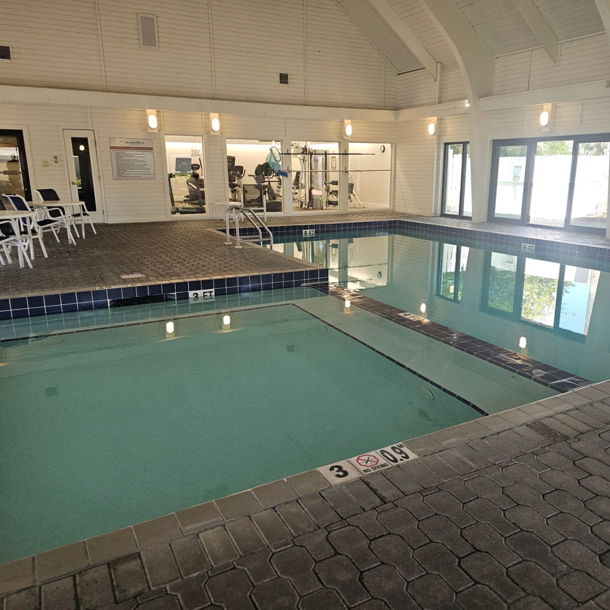 indoor pool with a fitness center in the back area along with tables and chairs on the pool deck