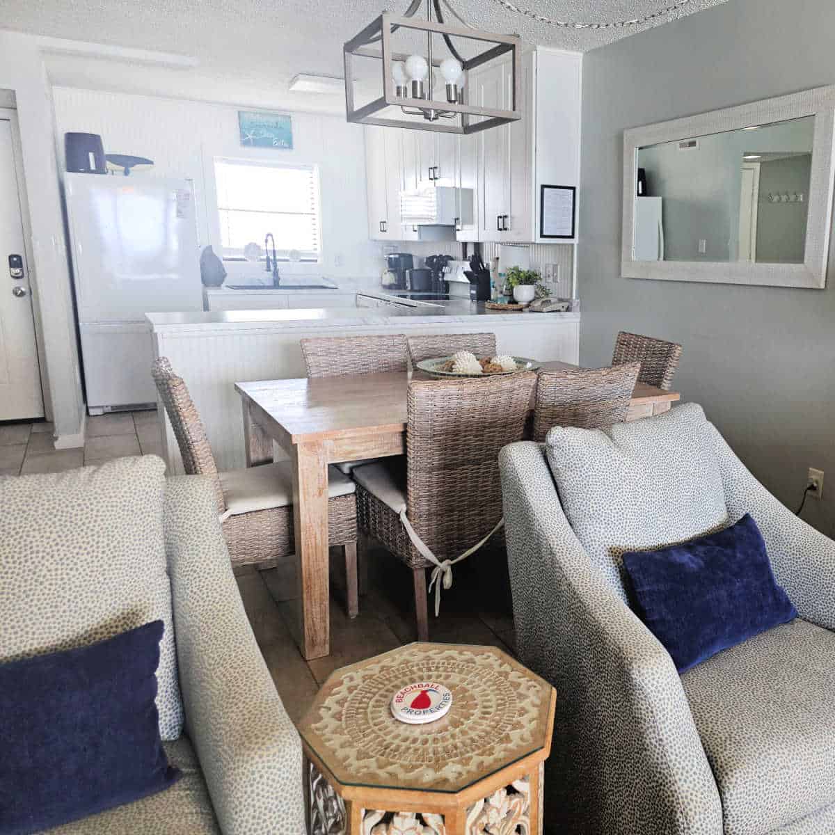 living room area with two chairs, a wooden table with Beachball Properties coasters, dining room table with 6 chairs, looking back to the white kitchen with light coming through the window
