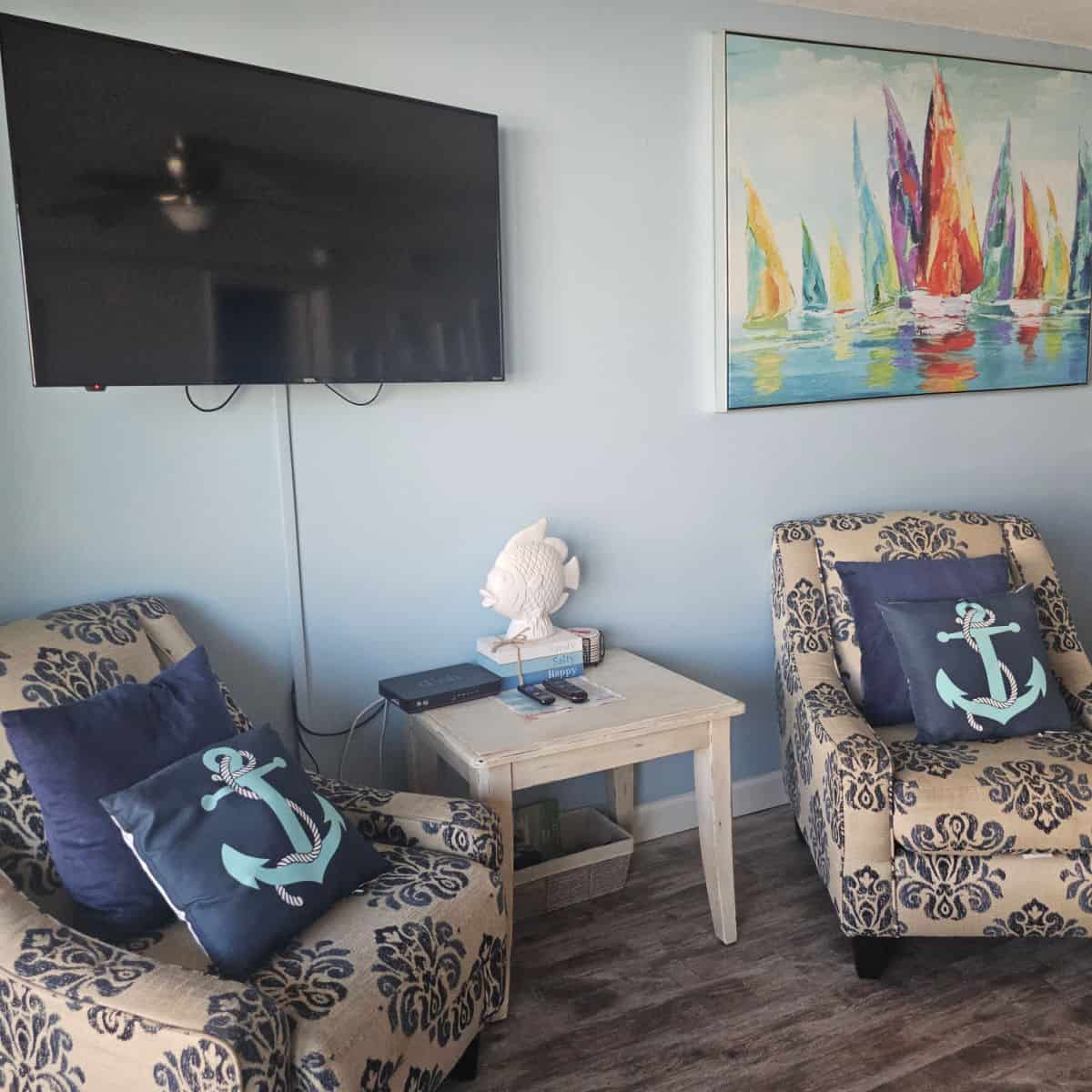 living room with a sailboat painting and flatscreen tv on blue walls, Two padded chairs wtih blue pillows and an anchor pillow, with a white table in between with books and a fish on it.