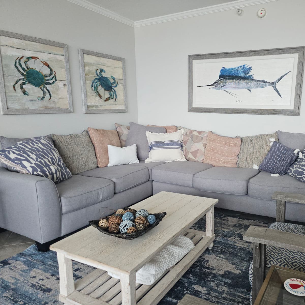 living room with grey couch covered in pillows, crab and marlin art on the wall, table in the middle of the carpet