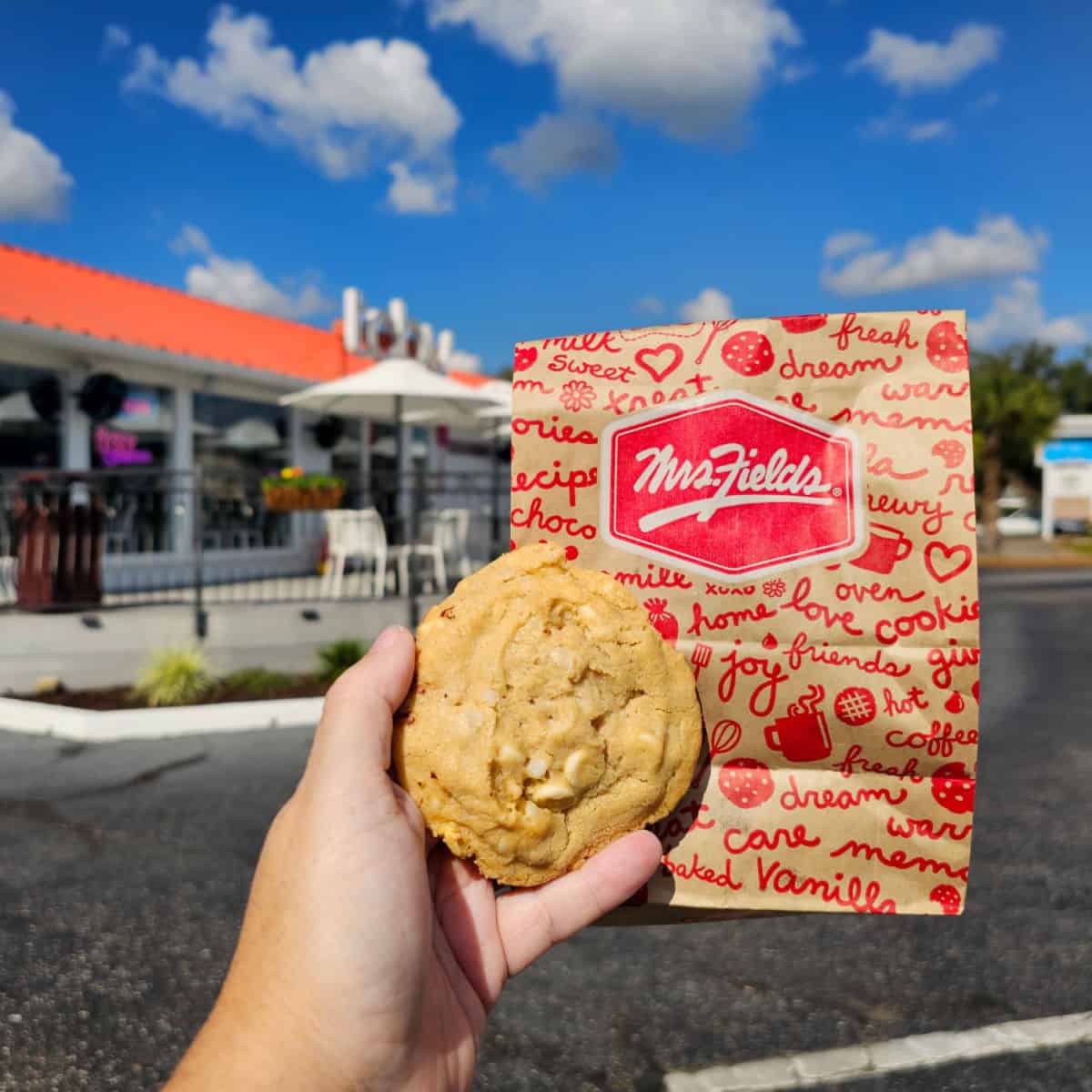 hand holding a cookie and Mrs. Fields cookie bag with building in the background