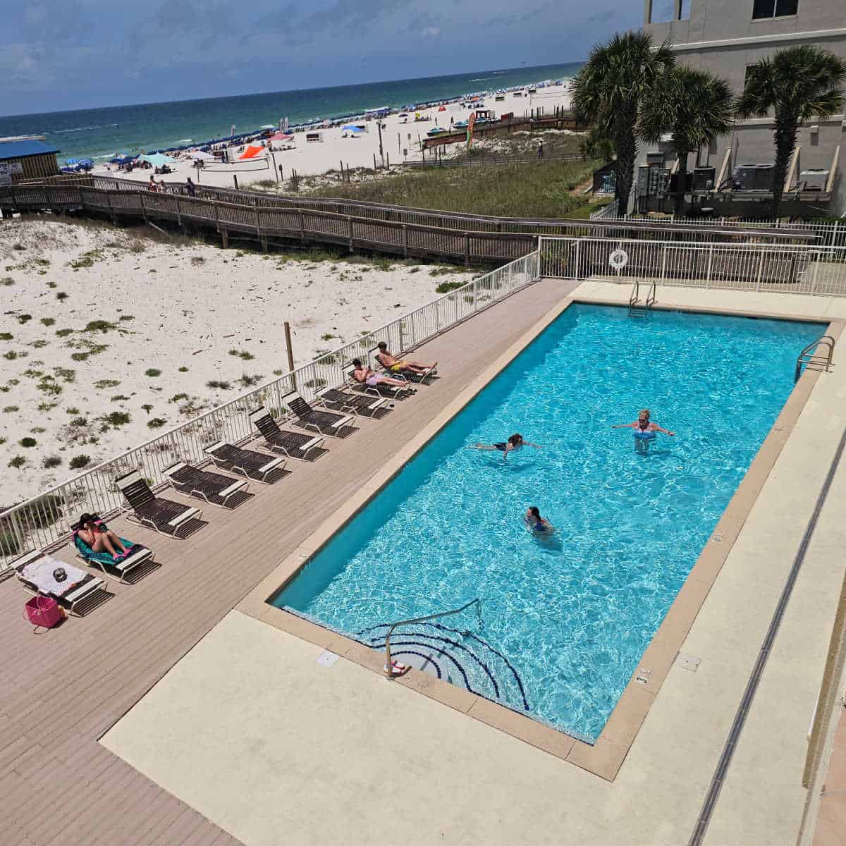 outdoor pool with people in the water, lounge chairs along the pool deck, boardwalk to Sea n Suds restaurant and the beach in the background.