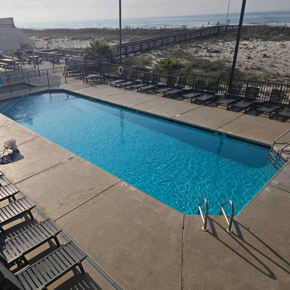 outdoor pool surrounded by deck chairs, fence around the pool aea, boardwalk over sand dunes