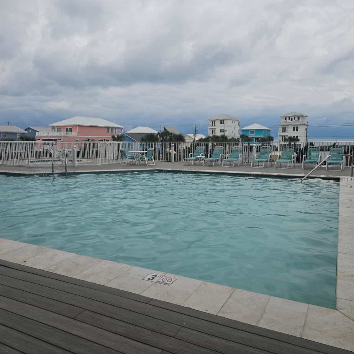 outdoor pool with lounge chairs around the deck, fence around the pool area, beach houses in the background