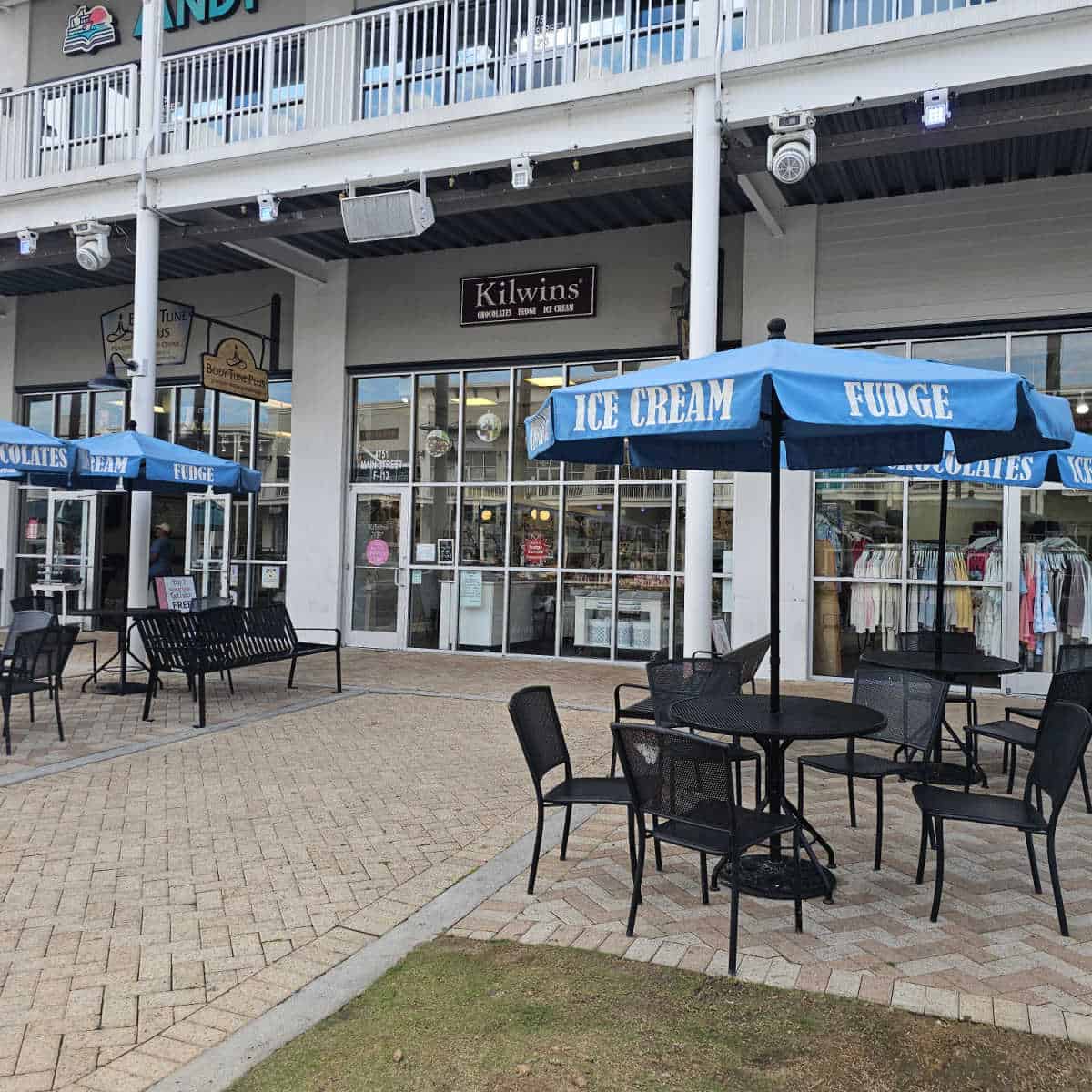 outdoor seating near the Kilwins store entrance with black chairs, black tables, blue umbrellas with ice cream and fudge printed on them.