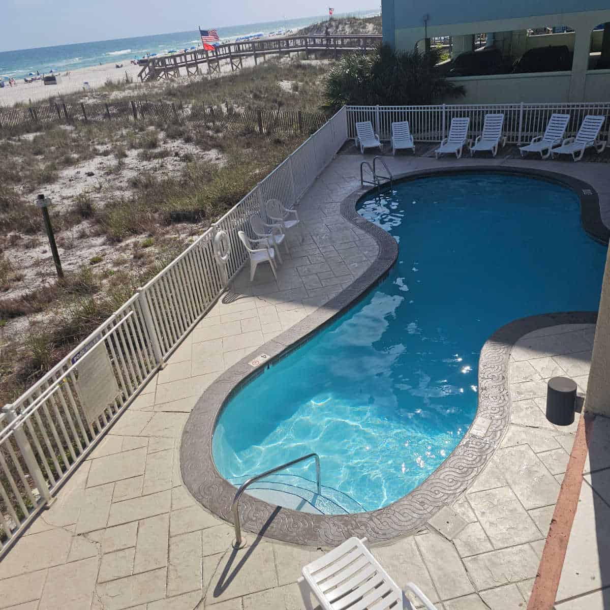 Looking down to an outdoor pool with white lounge chairs and chairs around it, fenced off from sand dunes with the Gulf of Mexico in the background.