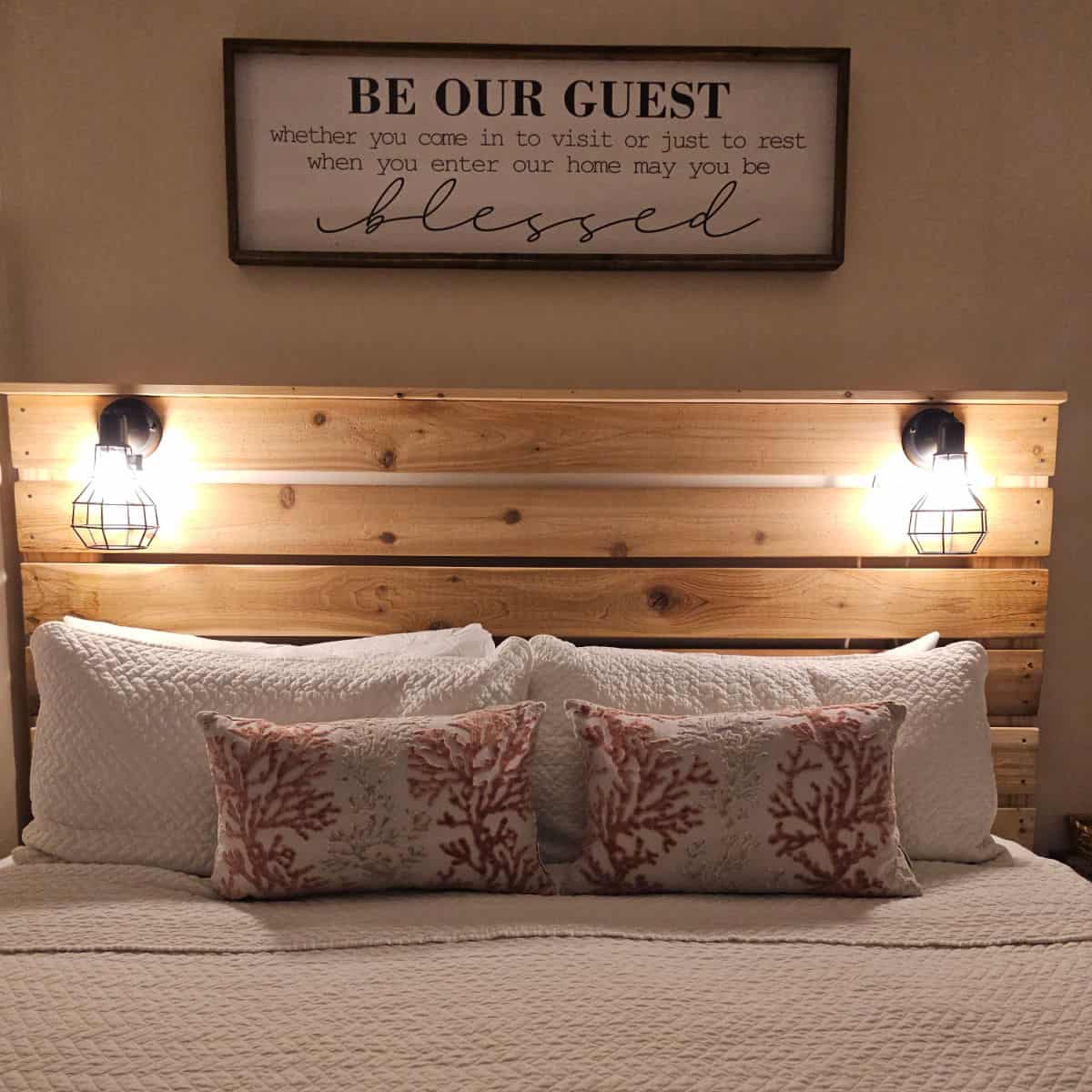 Be our guest art over a wooden headboard with lights and pillows on te bed.