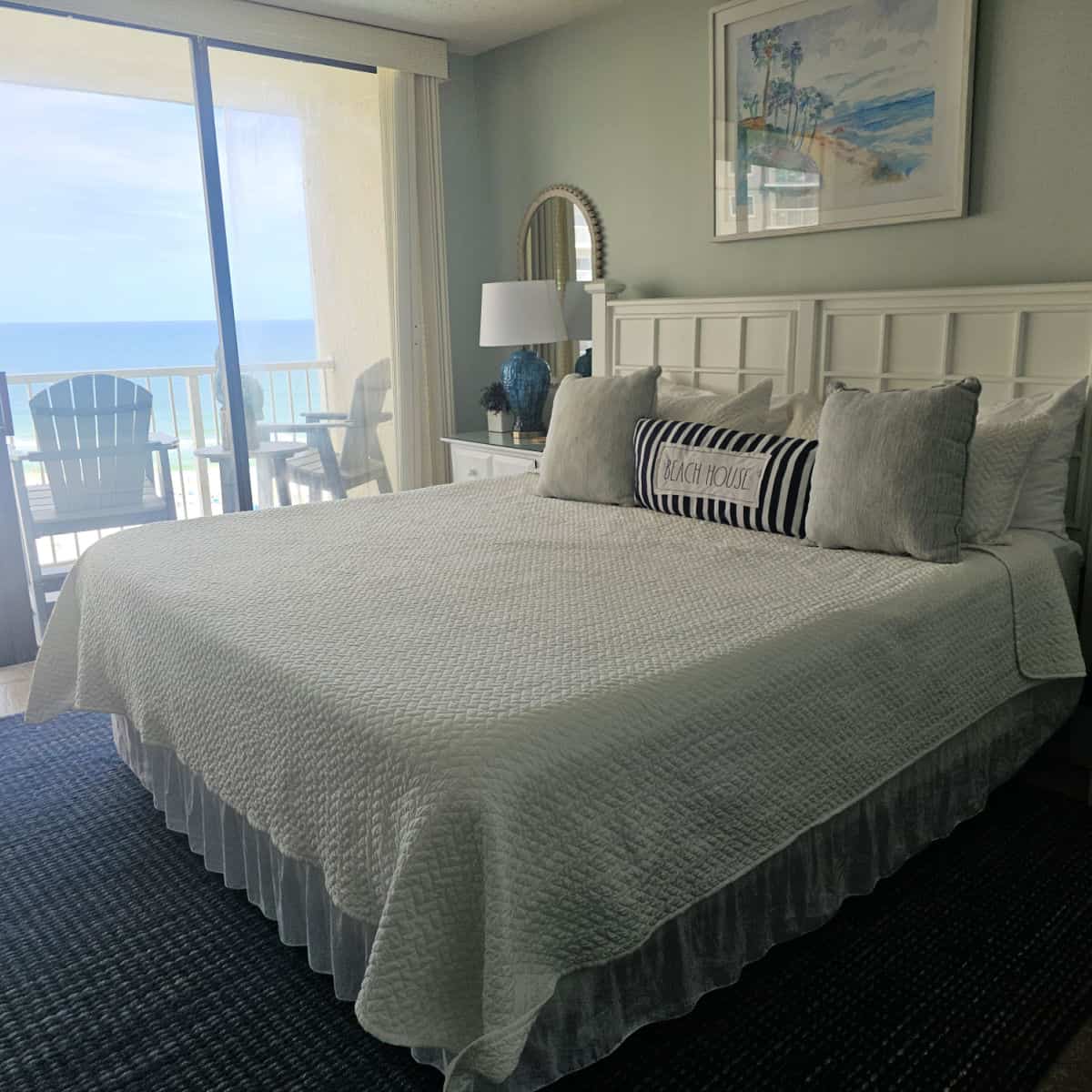 bedroom with king size bed with white linens, wooden headboard, beach scene art on the wall above the bed, nightstand with blue and white lamp, glass doors looking out to the Gulf with two Adirondack chairs and a table