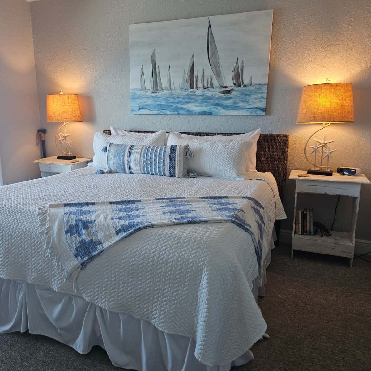 primary bedroom with large bed, white linens and pillows, sailboat art on the wall, two lamps on the side with nightstands