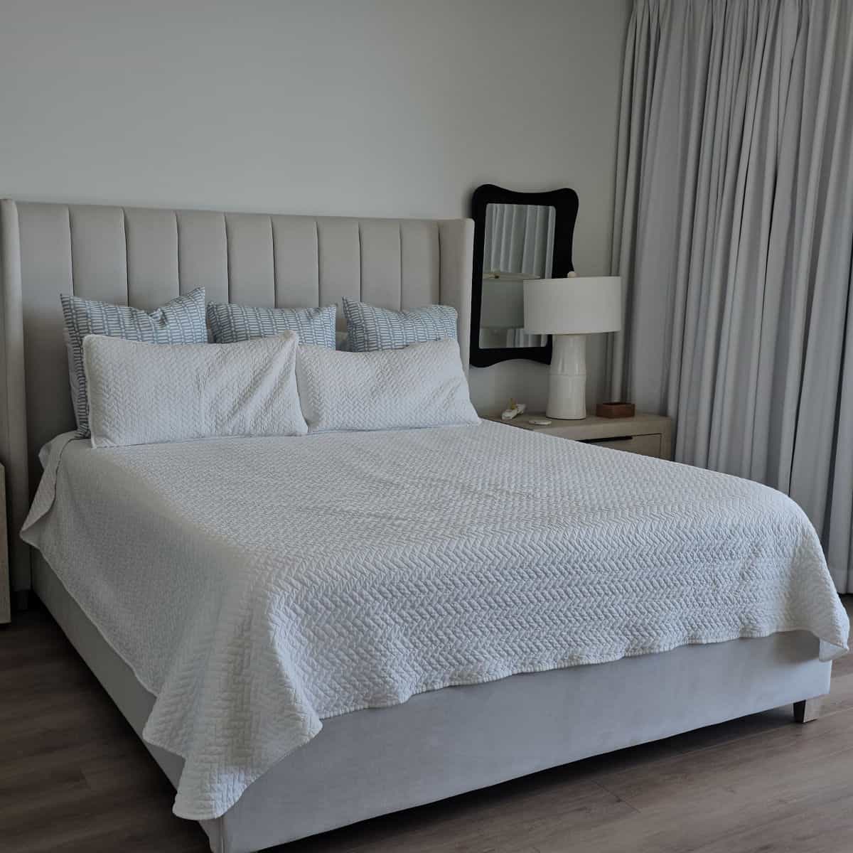 White bed linens on a king bed with a nightstand and white lamp, black mirror on the wall