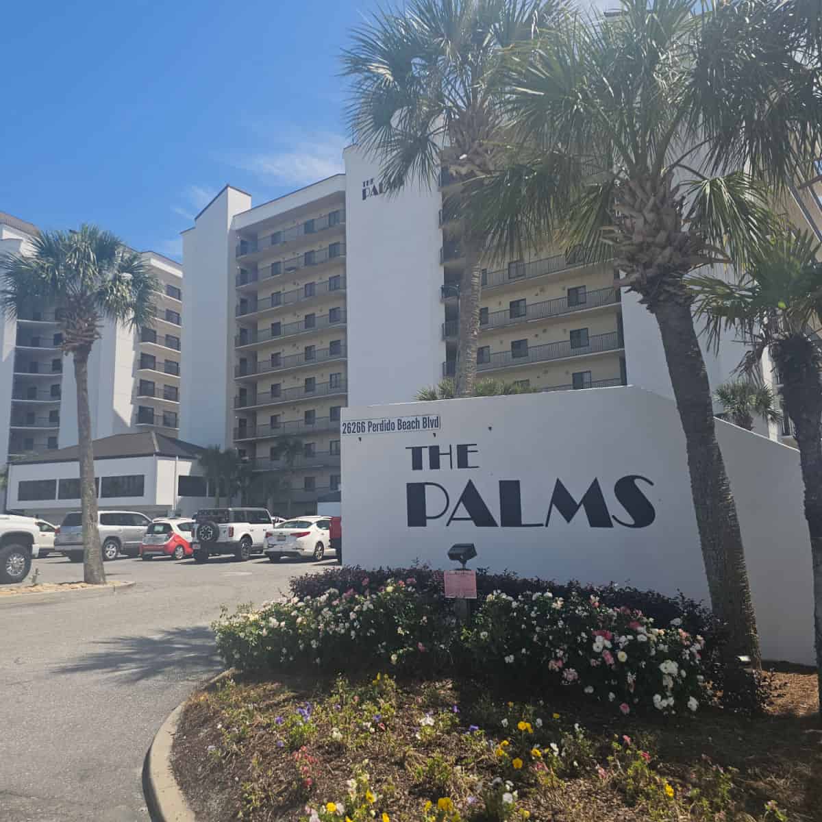 The Palms printed on a white sign, large build in the background with palm trees