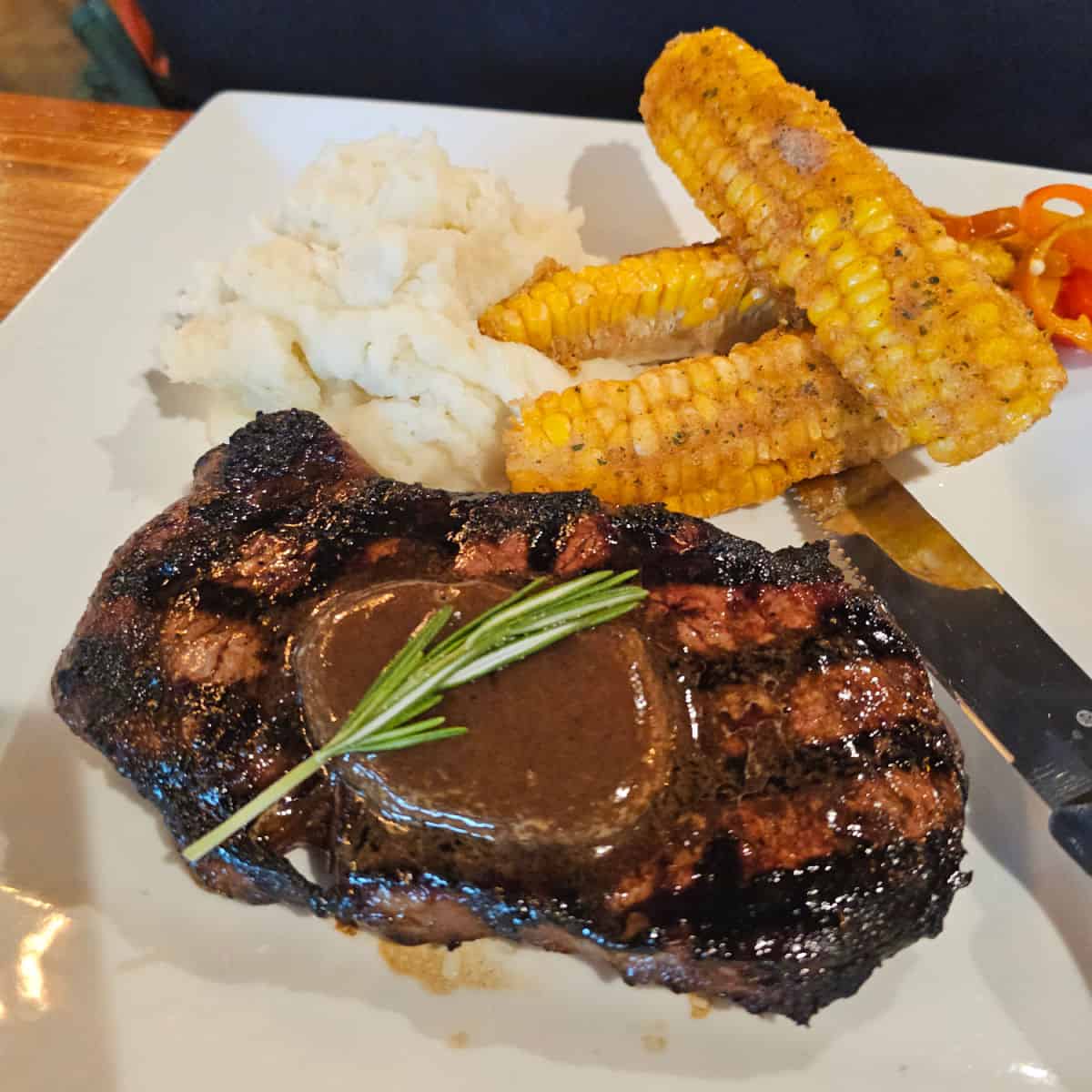 Steak with sauce and rosemary next to elote corn and garlic mashed potatoes on a white plate