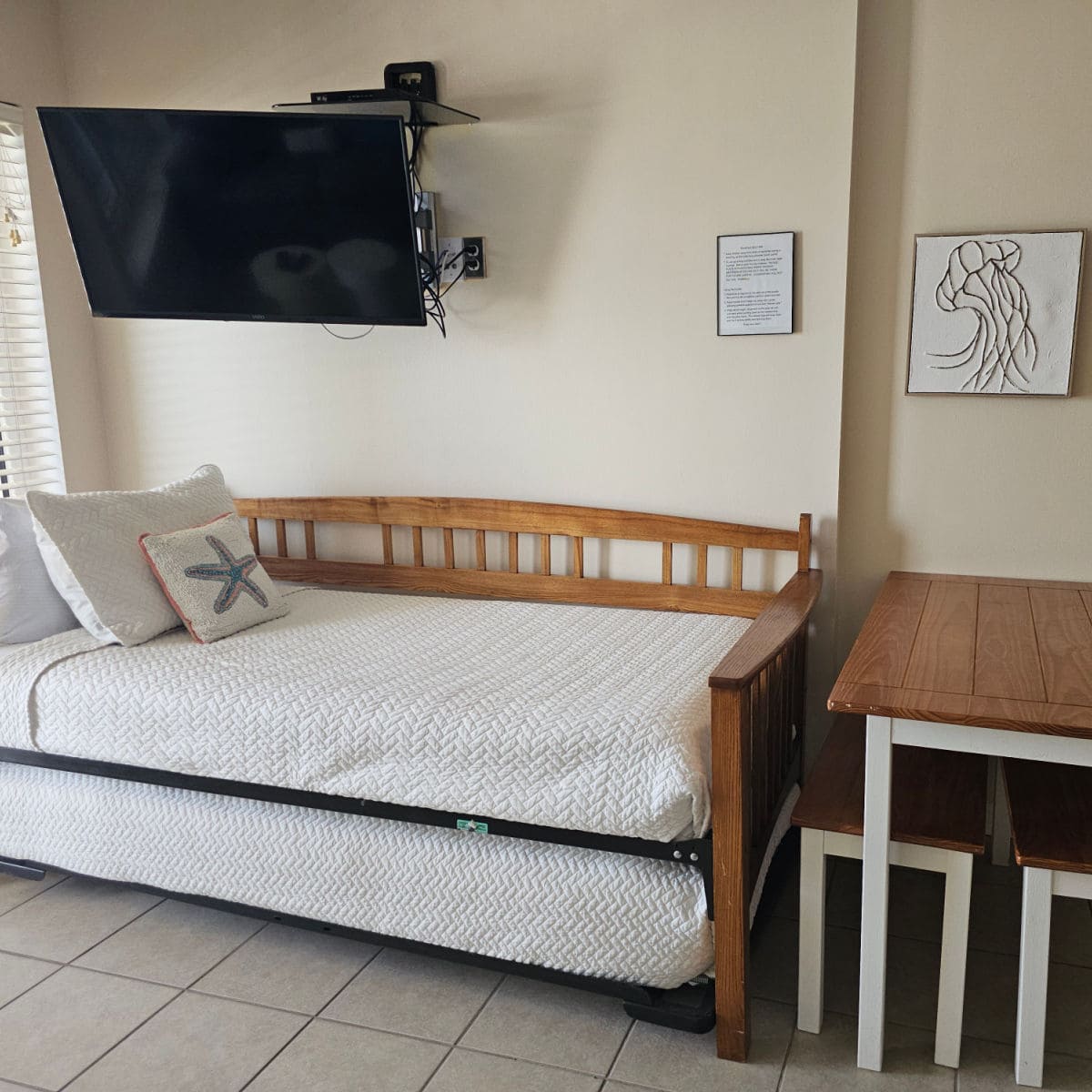 trundle bed with pillows under a flatscren tv, dining room table next to trundle bed