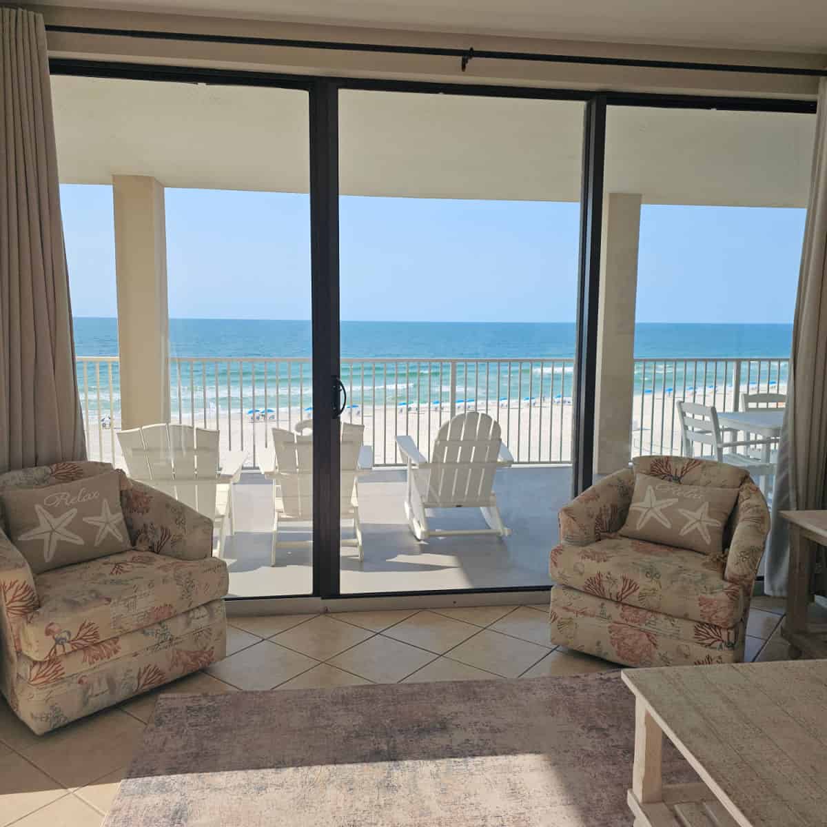 living room with two chairs with shell pillows and a deck with white Adirondack chairs gulf in the background and beach