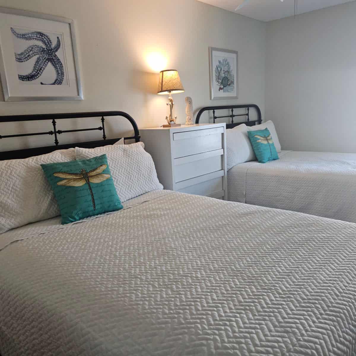 Two beds with a dresser in between them, white linens, pillows with dragonflies on them, coastal art on the wall