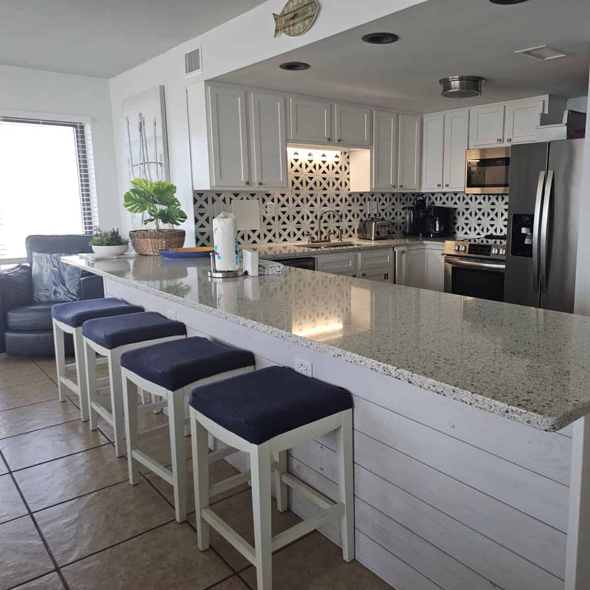 Kithen area with long counter, stools under the counter, blue chair in the corner, kitchen has stainless steel appliances and a gorgeous black and white backsplash