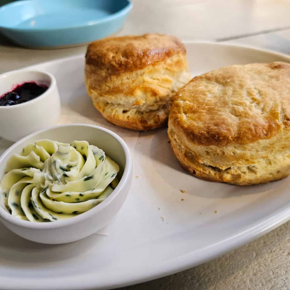 white platter with a bowl of herb butter, bowl of jam, and two large buttermilk biscuits