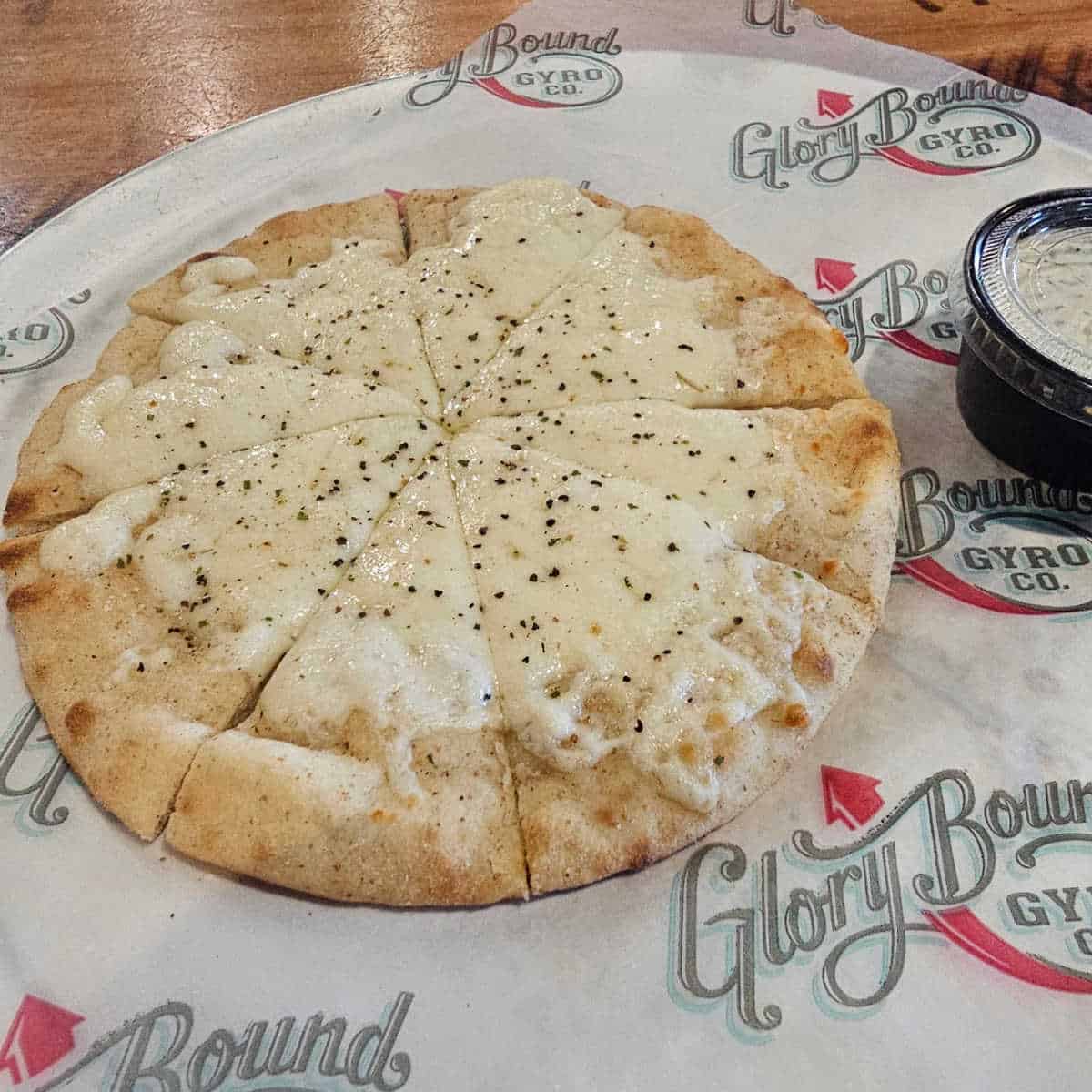 cheese covered pita bread on a glory bound gryo co paper lined plate with a container of dipping sauce