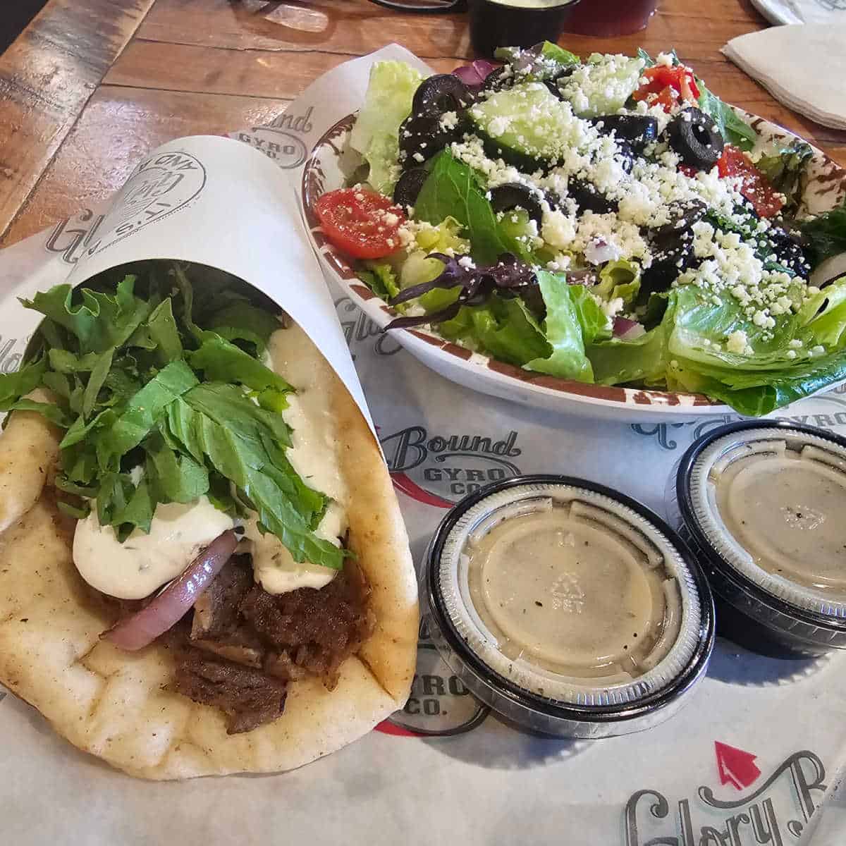 greek salad in a white bowl next to a gyro on a glory bound gyro co paper lined tray