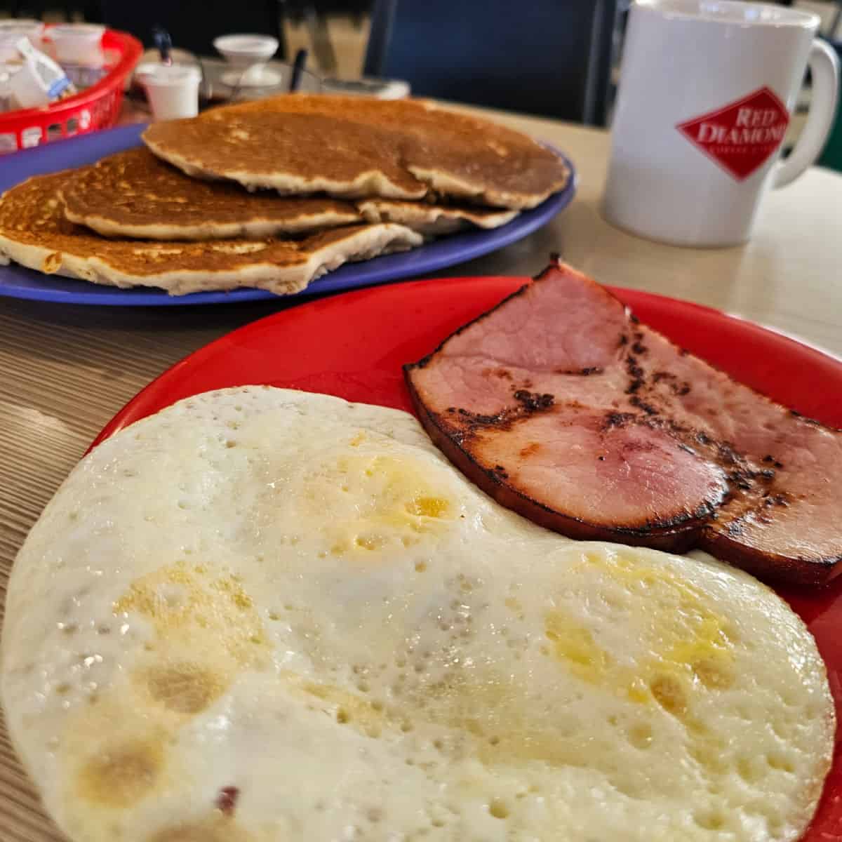 Ham and eggs on a red plate pancakes on a blue plate behind it
