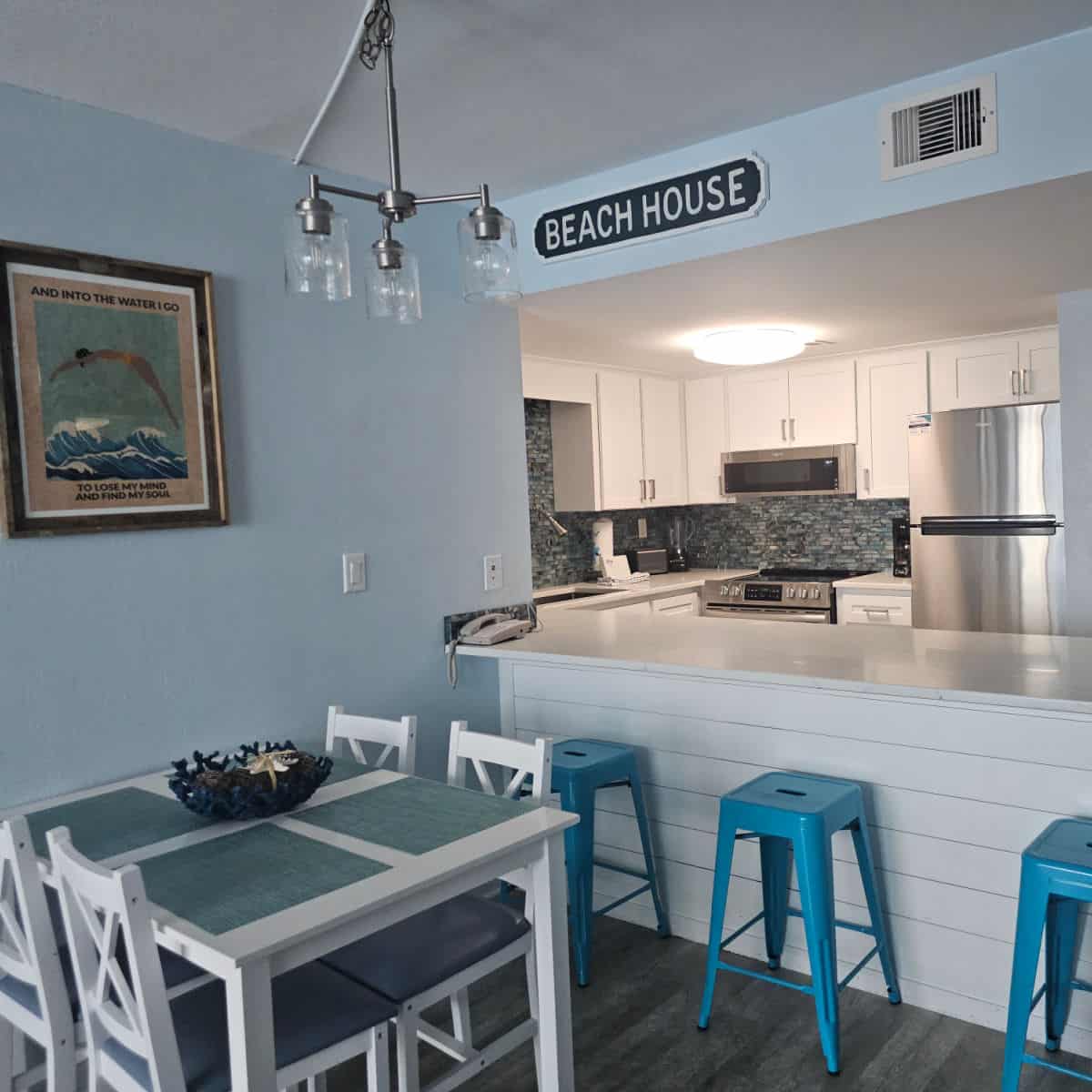 blue wall with art, small dining room table, blue stools at the counter with a small kitchen in the background.